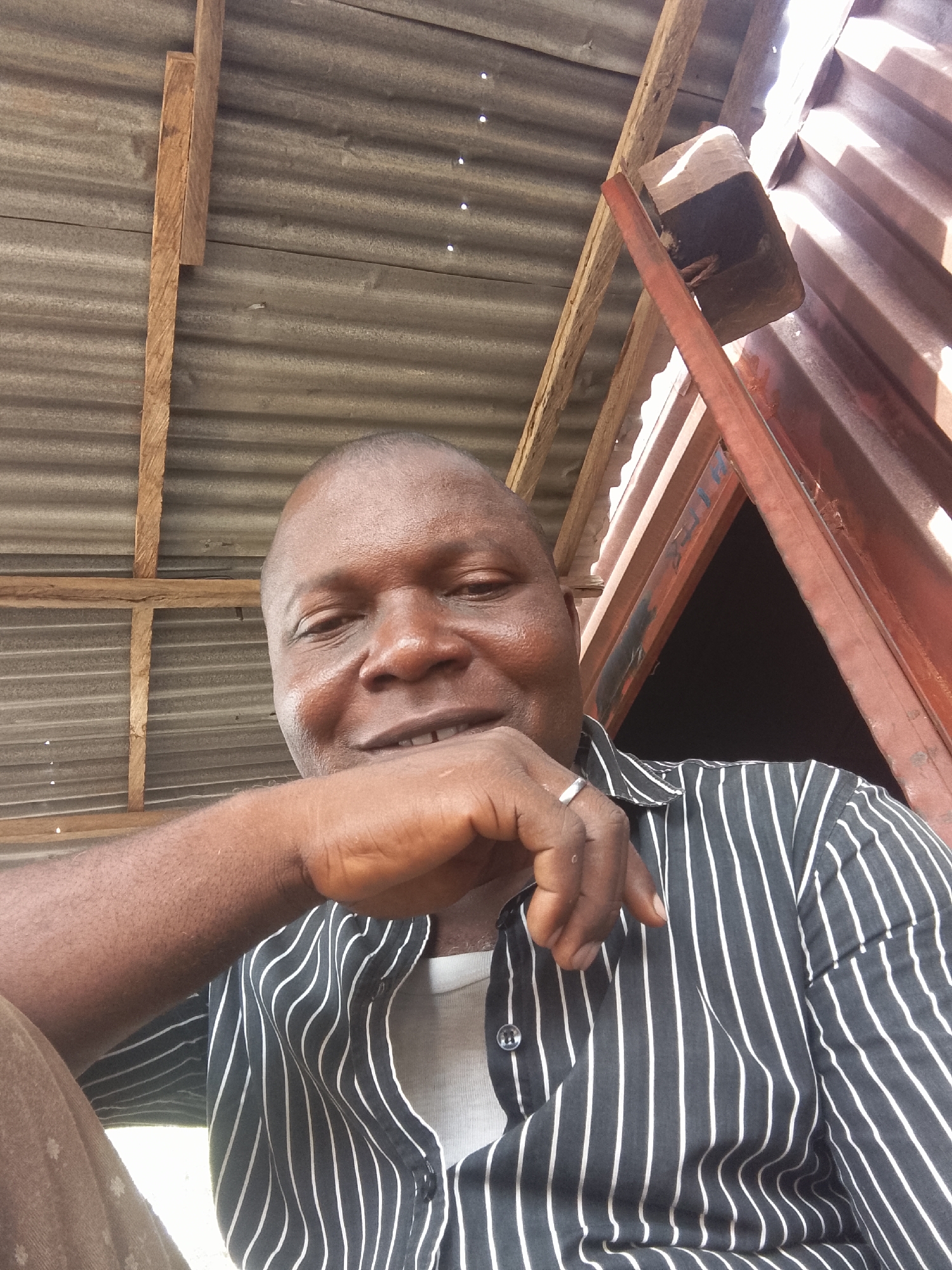 The image shows a close-up of a dark-skinned man with a shaved head. He is wearing a black and white striped shirt, with a white undershirt visible. His right hand is raised to his face, with a silver ring on one of his fingers. He appears to be smiling slightly.
The background consists of corrugated metal roofing and wooden supports. A reddish-brown metal structure with a dark opening behind it is also visible. The location is Unknown, Unknown.