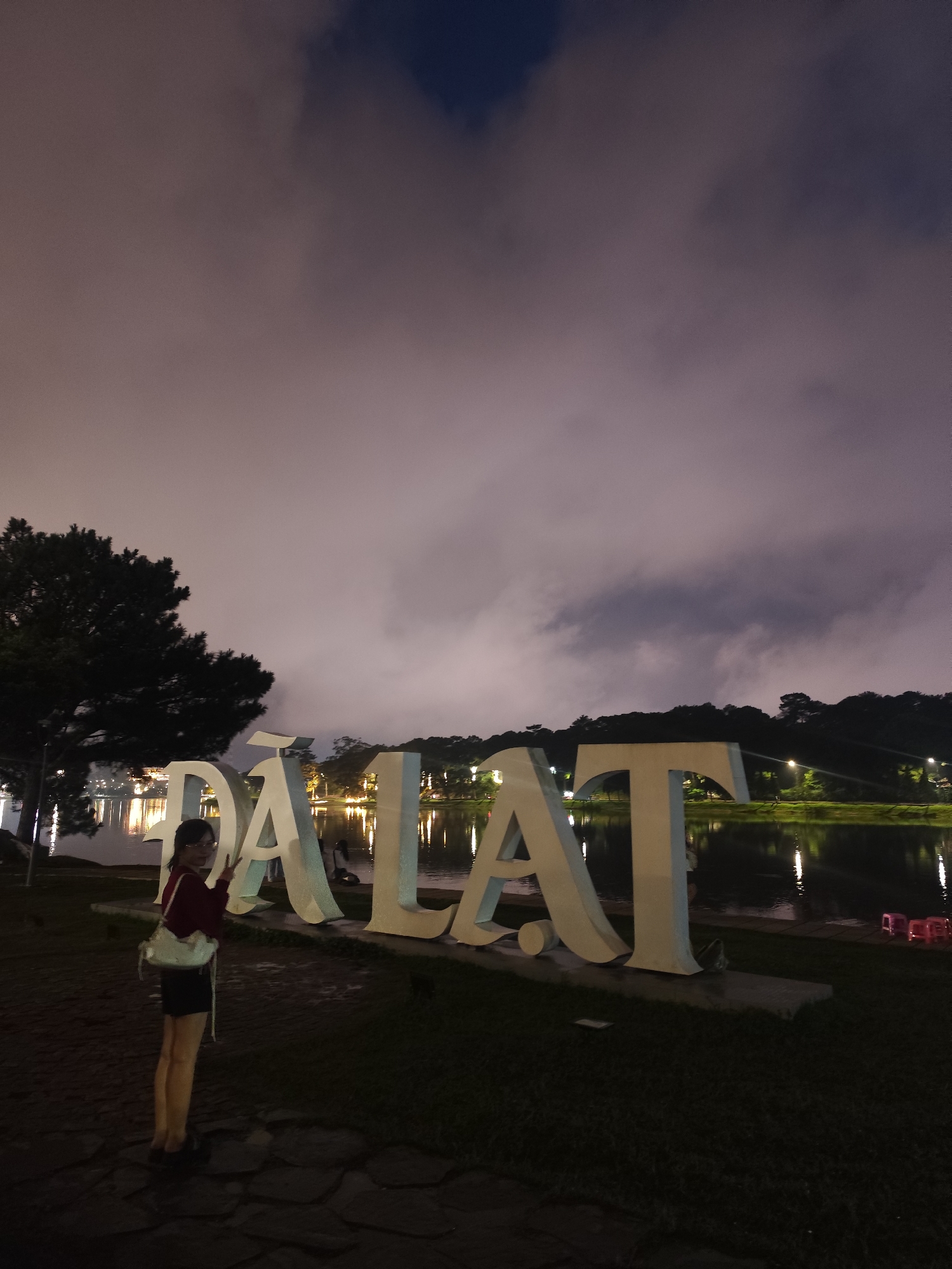 An individual, identified as female, is standing on a paved path in front of large, white, three-dimensional letters spelling "ĐÀ LẠT" at night. The individual is wearing a dark red top, dark shorts, and a white bag slung over their shoulder, with glasses on their face. They are looking towards the camera and holding up two fingers in a peace sign. Behind the letters, a body of water, reflecting lights from the opposite shore, is visible. The shoreline is lined with trees and more lights from a city or park. The sky is dark with clouds. The scene appears to be at Xuan Huong Lake in Da Lat, Vietnam, a popular tourist destination.