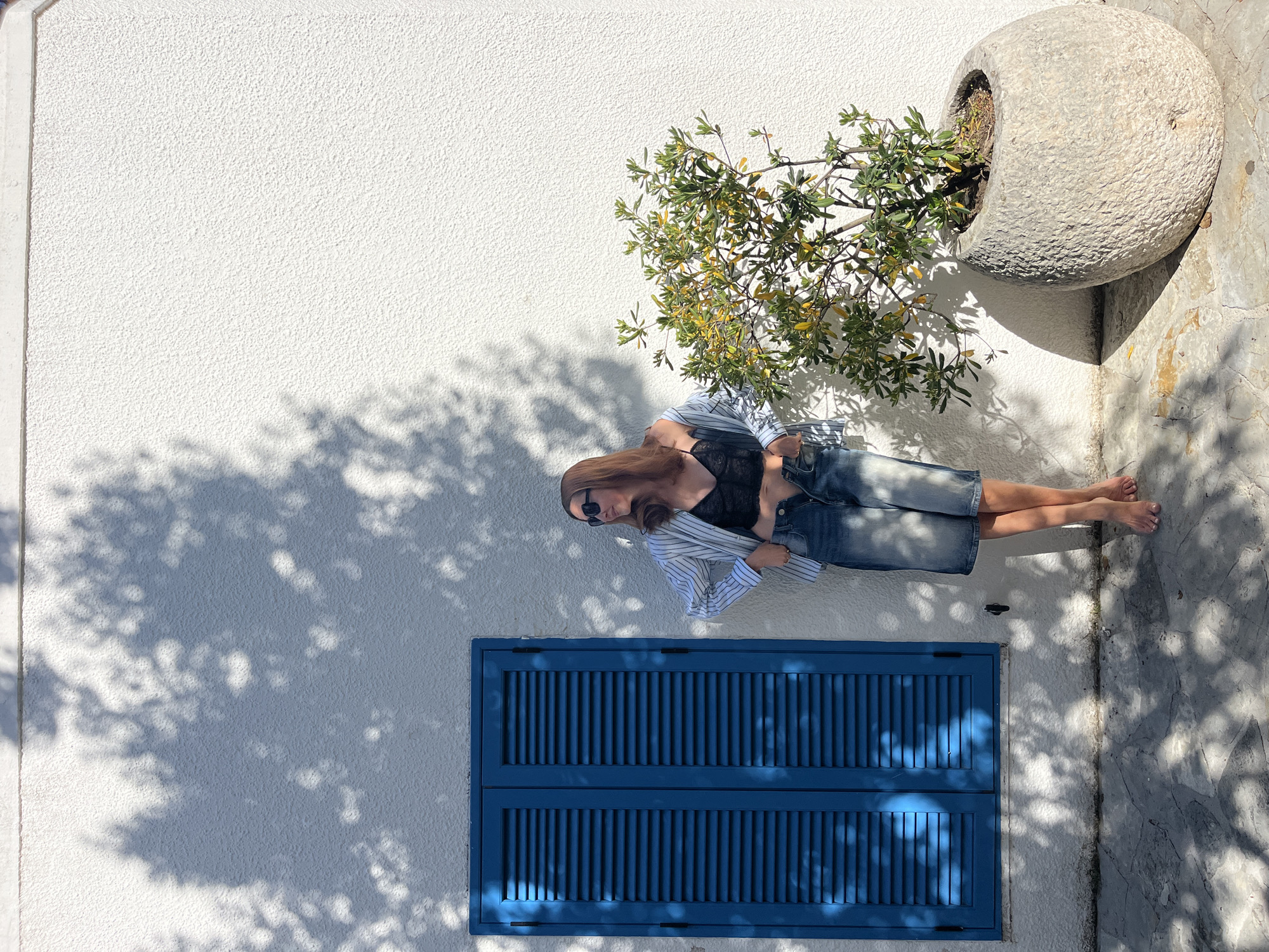 The image portrays a woman standing against a textured white wall in Budva, Montenegro, under bright, clear sunlight. The scene is captured from an elevated perspective, looking down onto the subject and surroundings.
The central figure is a woman with long, light brown hair, wearing dark sunglasses, a black top, and an unbuttoned light blue and white striped shirt over denim shorts. She is barefoot, with her feet resting on a light-colored, textured stone or concrete ground. Her posture appears relaxed, suggesting she is posing or taking a momentary break.
To the woman's left, a vibrant blue louvered window shutter is prominent against the white wall, adding a strong color accent. Above and slightly to her right, a large, rustic stone planter holds a green leafy plant with some yellowing leaves, contributing to the natural elements of the scene.
Sharp, intricate shadows of tree leaves are cast across the white wall and the ground, indicating a sunny day, likely mid-morning or early afternoon. The interplay of light and shadow highlights the textures of the wall and the ground. The clean white architecture, blue shutter, and natural stone planter evoke a quintessential Mediterranean setting. There is no visible text in the image.