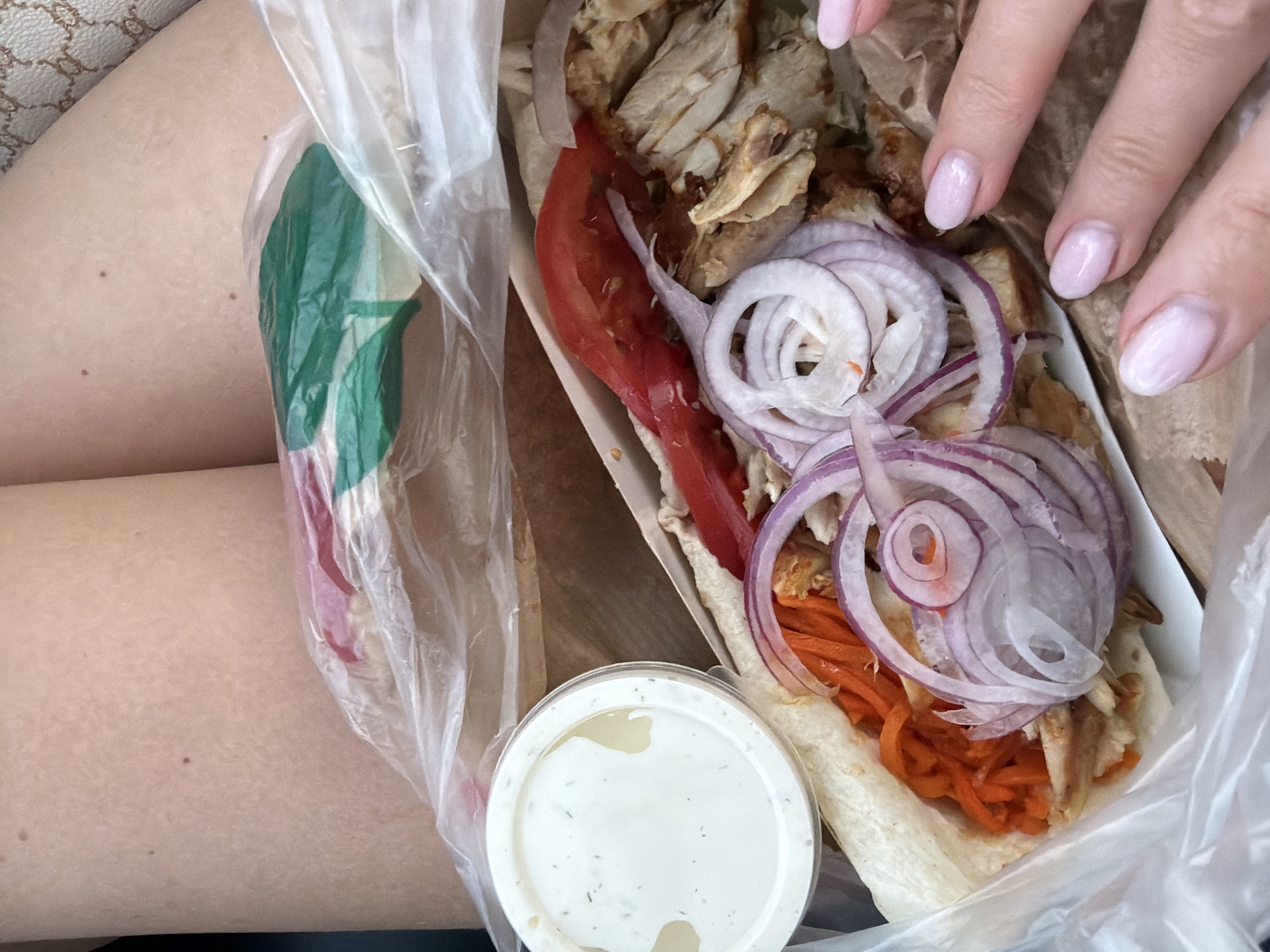 A top-down medium shot captures a person's legs and a takeaway food container filled with a shawarma. The shawarma, partially unwrapped from a white pita, reveals fillings of shredded chicken, julienned carrots, sliced red onions, and a slice of tomato. To the left of the shawarma is a small, clear plastic container holding a white sauce, likely a dipping sauce. The food is resting in a brown paper bag, with a clear plastic bag over the top. The person's bare legs, with visible moles, are in the upper portion of the frame, and a hand with light pink nail polish is reaching into the frame from the right. The background appears to be the interior of a car, with a patterned fabric visible on the seat. The scene is likely outdoors during the daytime, given the lighting. There is no specific indication of the city of Volzhskiy, Russia, in the image.