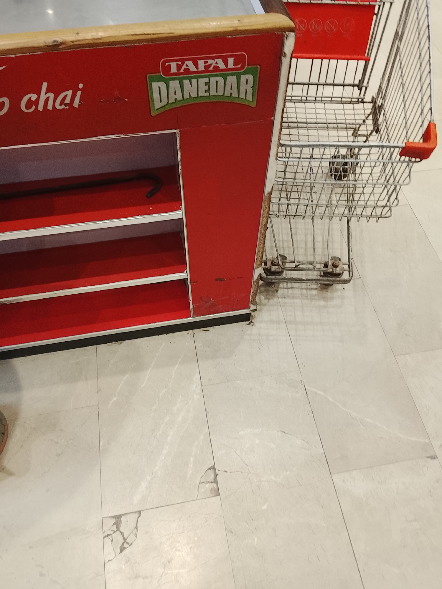 A retail shelf unit bearing the brand name "TAPAL DANEDAR" stands adjacent to a wire shopping cart in an indoor space with marble-like tiled flooring. The red shelf unit has several empty shelves. The shopping cart, composed of wire mesh, appears slightly used, with minor rust on the wheels. The location is inferred to be in Gulgasht Colony, Pakistan, based on the brand name commonly found there. No people are visible in the frame. The scene suggests a shop or a retail environment.