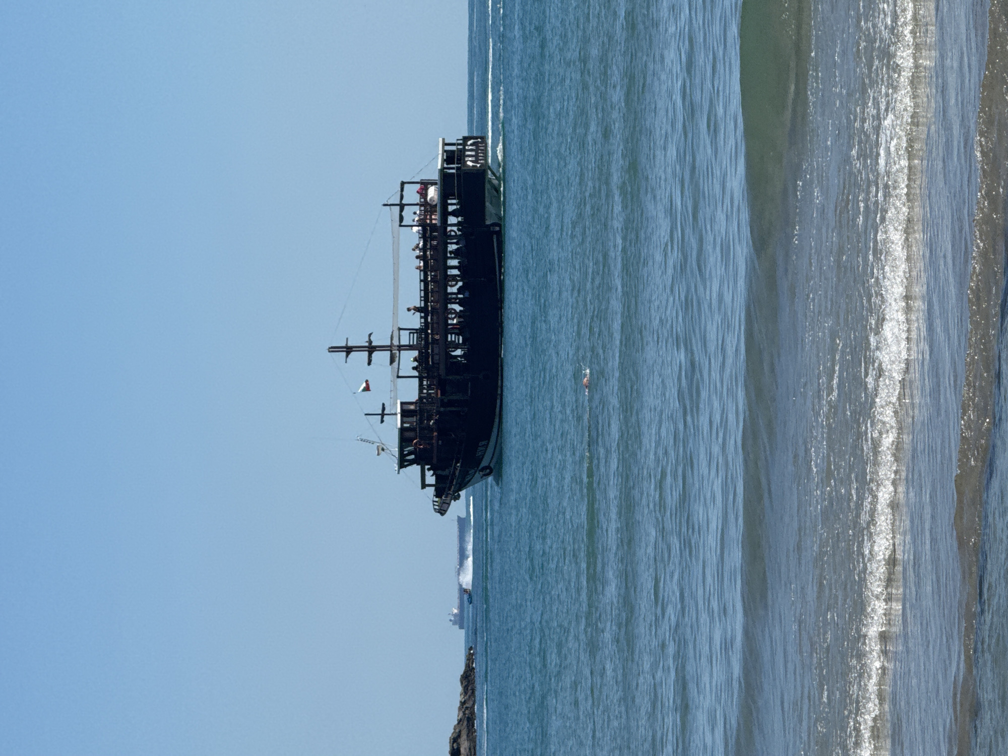 A black, multi-decked pirate-style tourist boat with a white awning and multiple masts is sailing on a bright blue sea under a clear sky. The boat has many people on its upper deck. A smaller boat is creating a wake behind it. In the distance, a larger cargo ship is also visible. In the foreground, gentle waves are approaching a shore, and a single person is swimming in the water. The scene appears to be near the coast of Sozopol, Bulgaria, with rocky outcrops visible to the left.