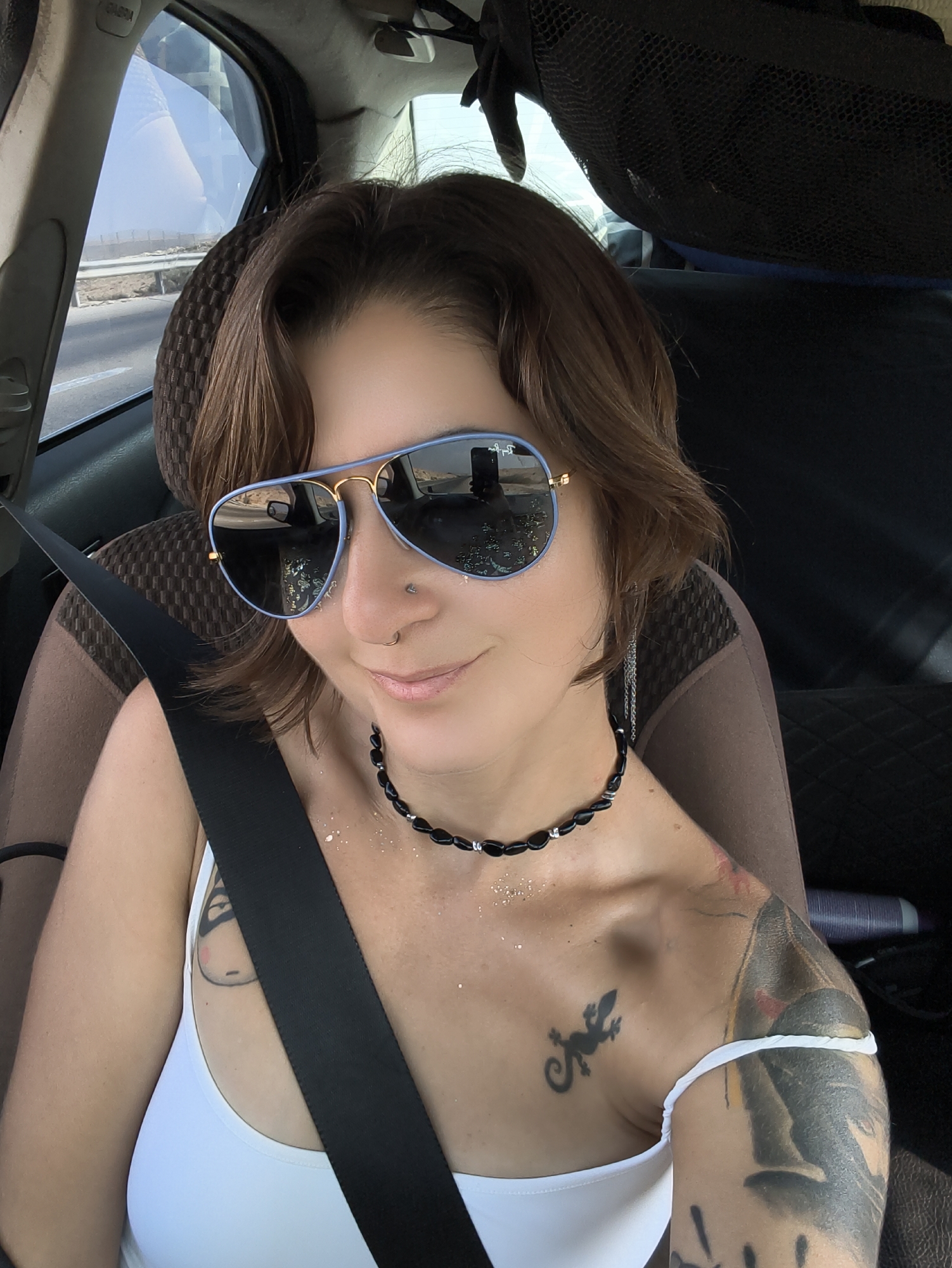 A woman with short, brown hair and sunglasses is sitting in the driver's seat of a car. She is wearing a white tank top and a black beaded necklace. A seatbelt is diagonally across her chest. There are tattoos visible on her arms and chest. The interior of the car appears to be a standard model, with a dark headrest and a black mesh organizer visible in the background. Sunlight streams in from the side window, illuminating the interior and casting shadows. The image appears to have been taken on a road, possibly in a desert landscape, given the arid environment visible through the windshield. The location is identified as the Ramat Negev Regional Council, Israel.