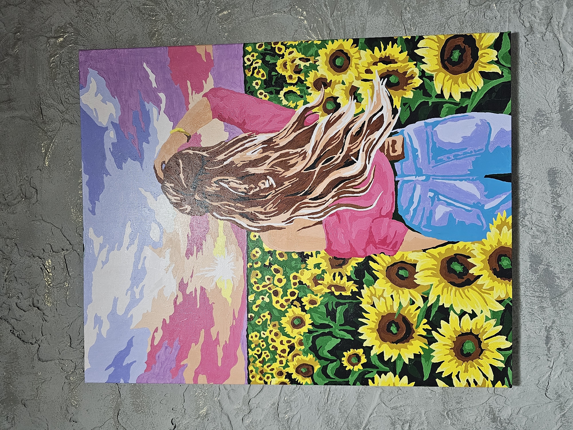 A painting on canvas depicts a woman from behind, standing in a field of sunflowers and looking towards a sunset. The woman has long, flowing brown hair and is wearing a pink off-the-shoulder top and light blue jeans. The sky is filled with clouds in shades of purple, pink, and white, with the sun setting in the distance. The sunflower field is dense, with vibrant yellow petals and dark brown centers. The painting is set against a textured gray wall, which is not part of the depicted scene. The image does not contain any information about the city of Kalinovka, Russia.