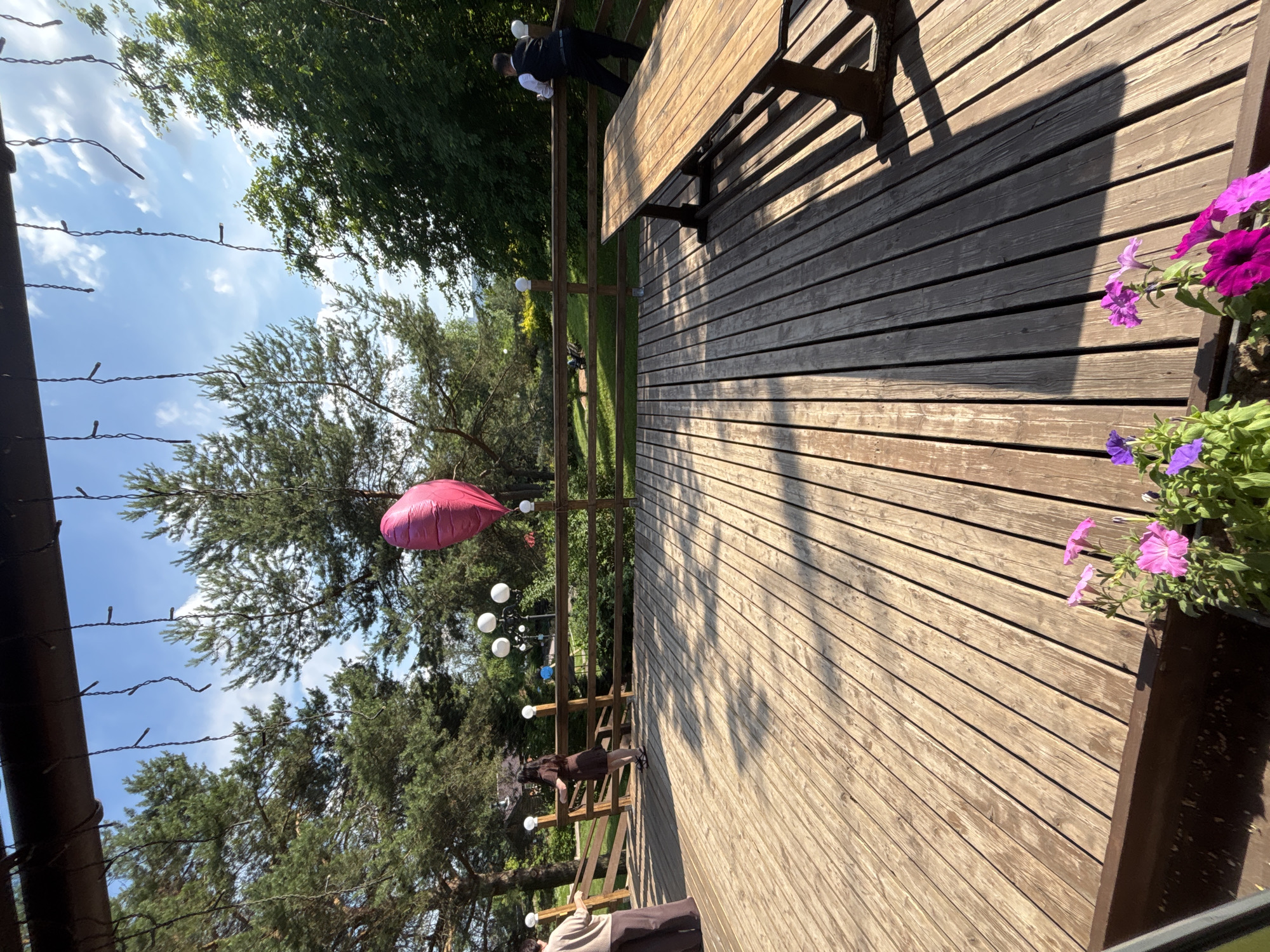 A spacious wooden deck is set up for an outdoor event, possibly a wedding reception or party, in Izhevsk, Russia. The deck is surrounded by a rustic wooden fence and overlooks a grassy area with trees. A large wooden picnic table and benches are positioned on the deck, suggesting a dining or gathering area. Several white spherical lights and a bright pink heart-shaped balloon are visible, adding a festive touch to the scene. Two women are standing on the deck, one in a beige top and brown pants, the other in a brown dress. A man dressed in a white shirt and black vest is leaning against the fence. The sun casts long shadows across the wooden planks, indicating it's late afternoon or early evening. Flower pots with pink and purple flowers are placed at the edge of the deck. The sky is blue with scattered white clouds, and lush green trees form a backdrop to the event space. The overall atmosphere is one of celebration and outdoor leisure.