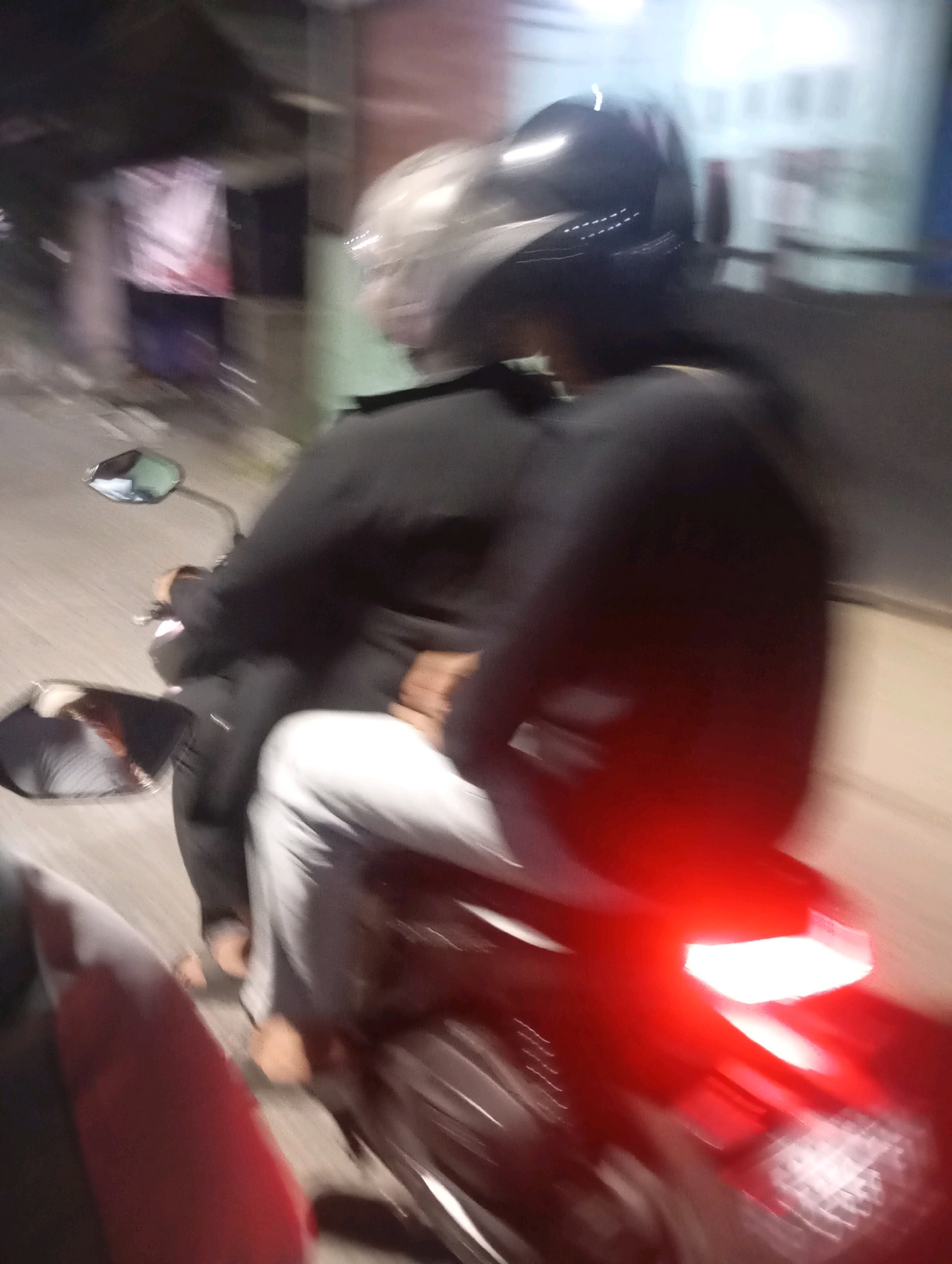 The image depicts two individuals riding a motorcycle at night. The motion blur suggests the motorcycle is in motion, likely on a street. The rider in front wears a dark helmet and a dark jacket. The passenger behind them wears a clear or light-colored helmet and a dark jacket. The passenger's left arm is visible, resting on the rider's jacket. The lower half of the image shows the motorcycle's tail light, which is illuminated red. The background is blurred, showing indistinct structures that suggest an urban or suburban environment. Based on the provided metadata, this scene is located in Babelan, Indonesia.