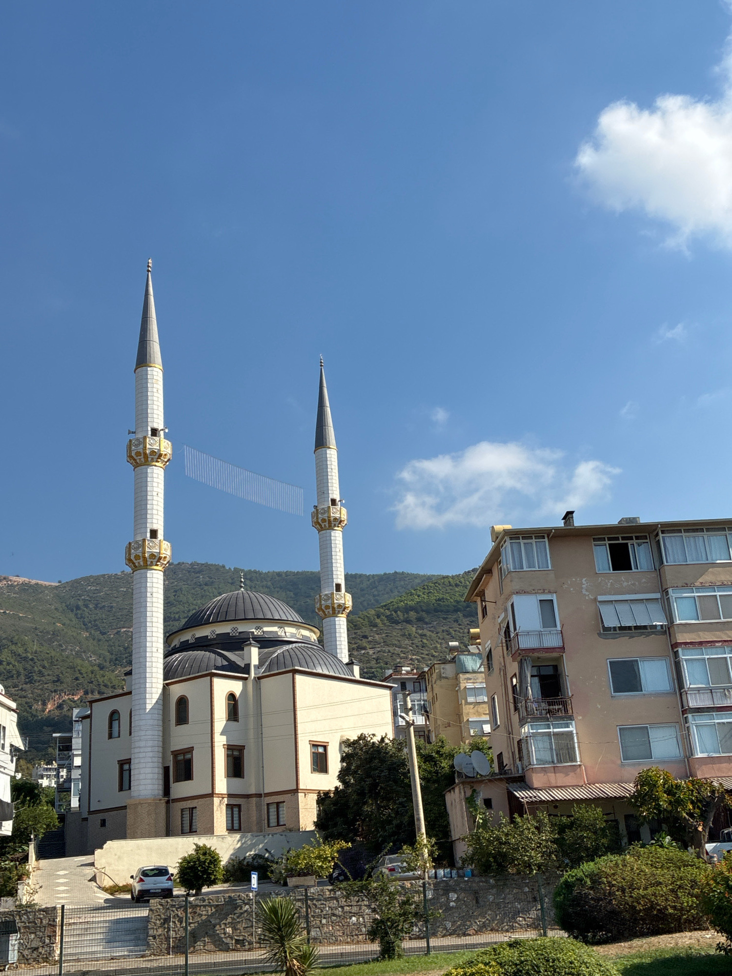 The image depicts a mosque with two prominent minarets, a central dome, and an accompanying building in Alanya, Turkey. Adjacent to the mosque, a multi-story residential building with balconies and numerous windows is visible. In the background, a tree-covered mountain slopes upwards. The sky is blue with a few scattered white clouds. A white car is parked on a paved road in front of the mosque. A stone wall and vegetation are in the foreground.