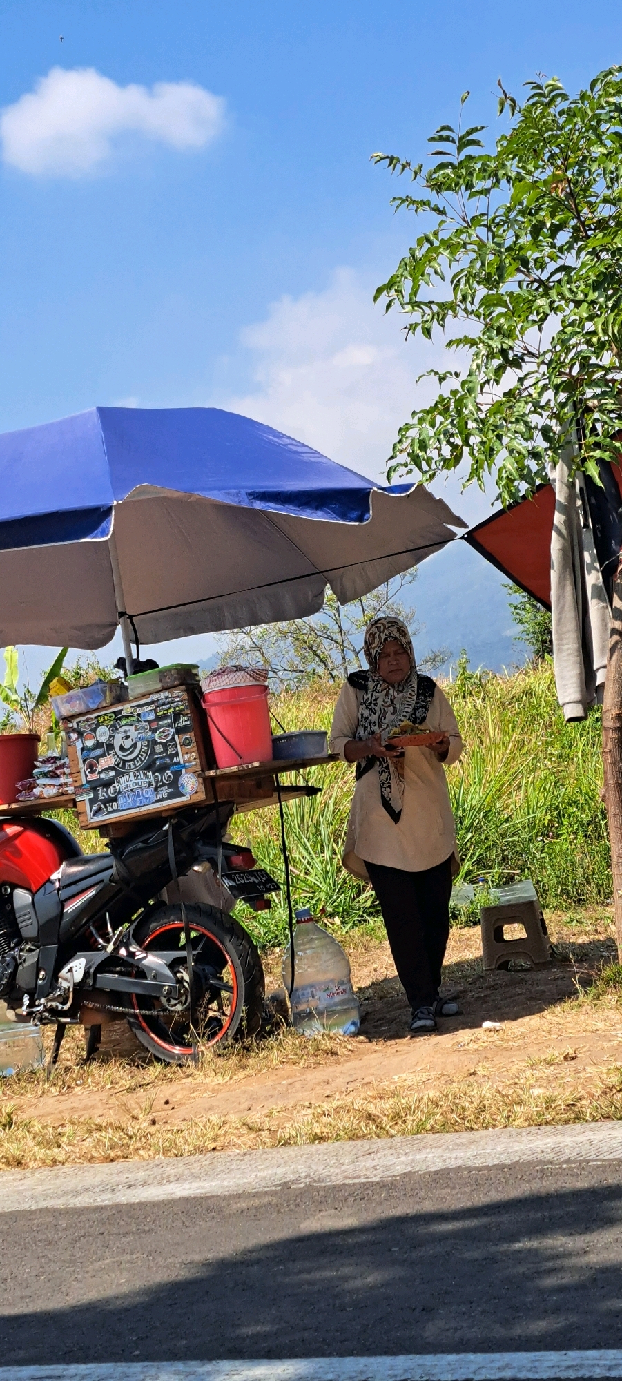 A woman in a headscarf and a light brown tunic stands holding a plate of food near a motorcycle-mounted food cart. The cart is covered by a large blue and grey umbrella. The woman is dressed in black pants and grey shoes. The cart is adorned with stickers and has two red buckets and a blue container on top. A large bottle of water is placed next to the motorcycle. The background shows a grassy area with trees and a distant mountain range under a clear blue sky with scattered clouds. The scene appears to be located along a roadside. The image was taken in Sekarjoho, Indonesia.