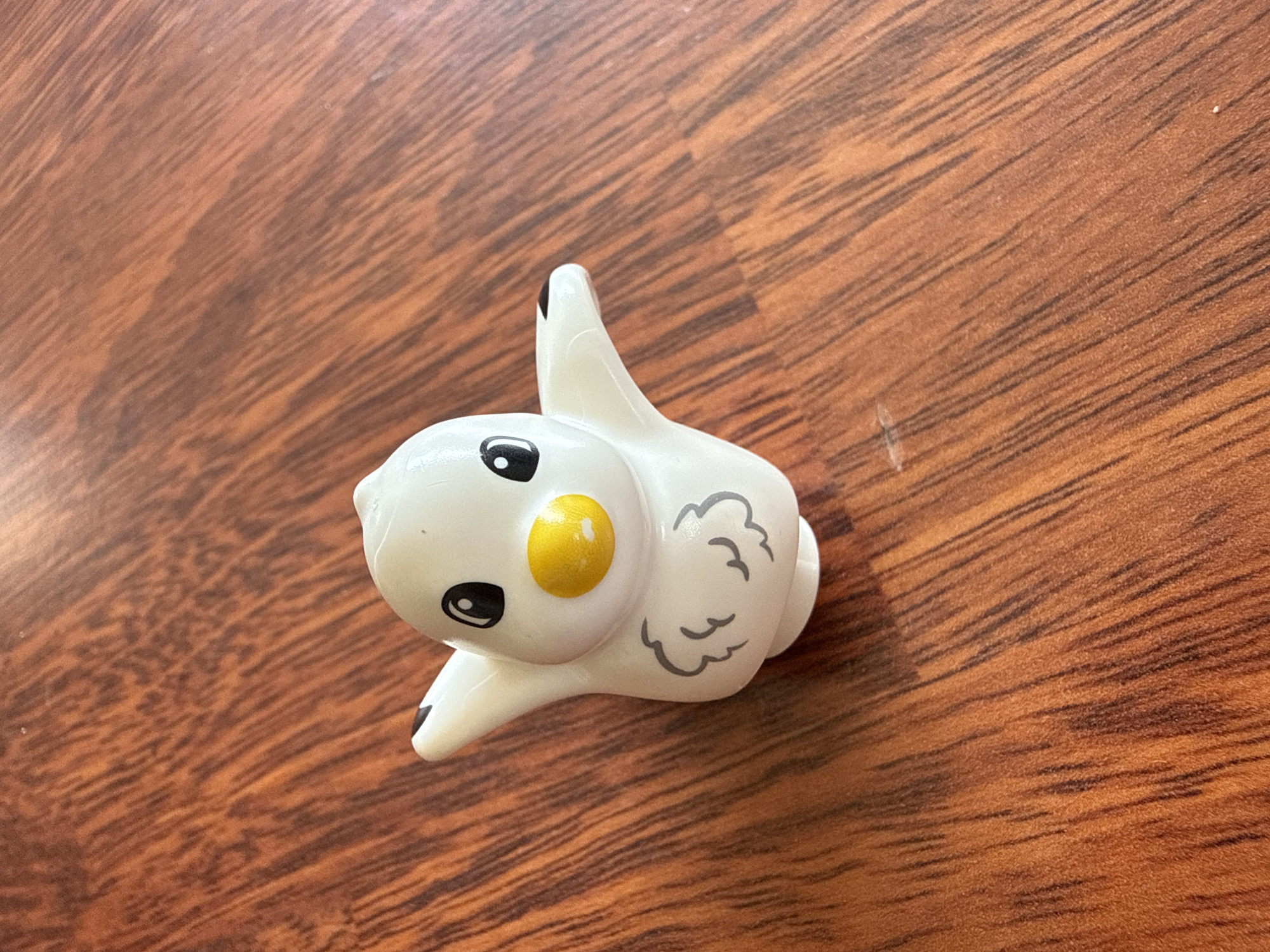 A white plastic duck toy with black eyes, a yellow beak, and grey feather markings on its chest sits on a brown wooden surface. The duck's arms are outstretched. The background is a wooden tabletop with visible grain patterns. The image is taken from a top-down perspective.