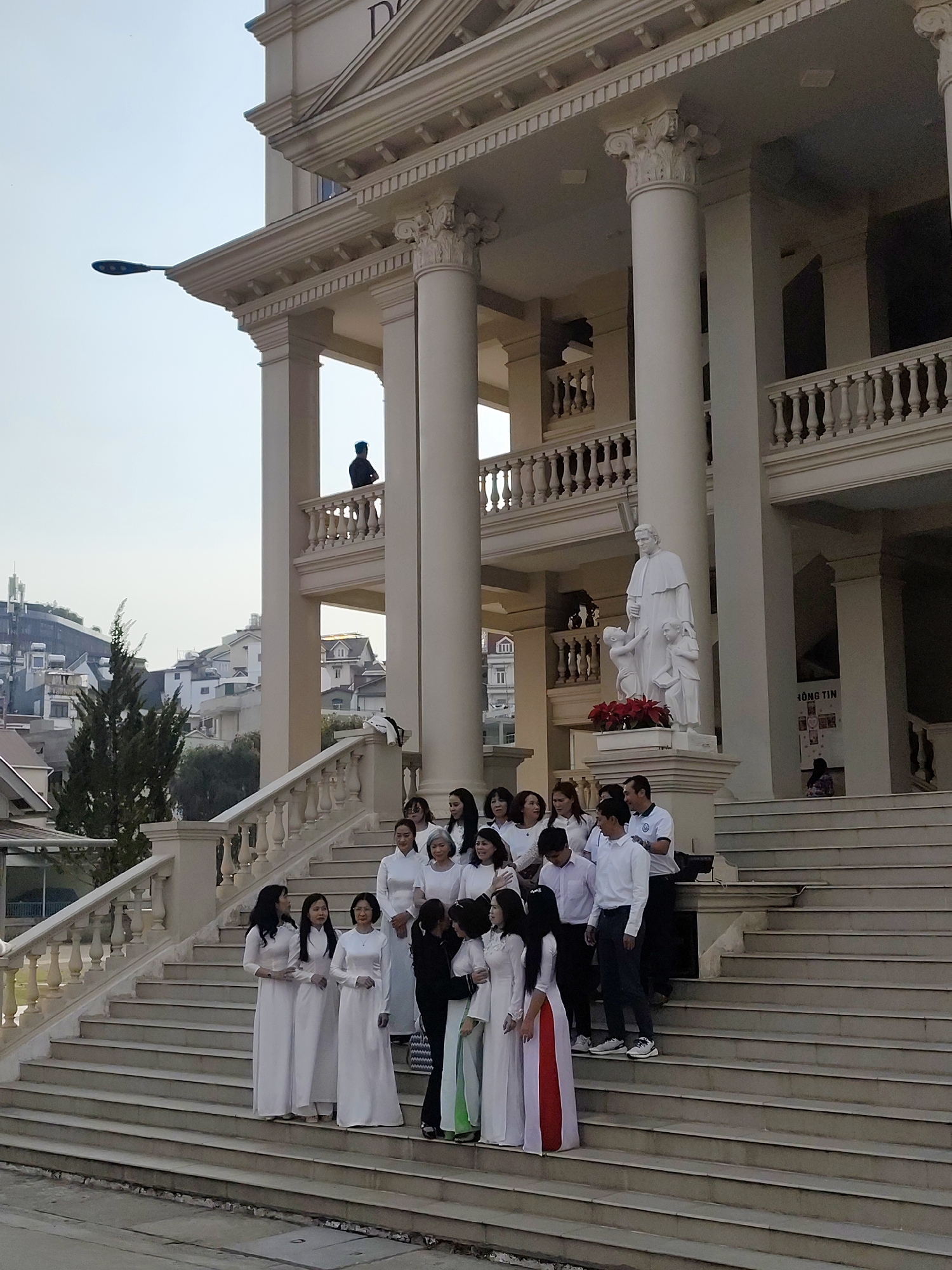The image shows a group of approximately 16 people posing on the steps of a large building, likely a school or institution, in Da Lat, Vietnam. Most of the women are wearing traditional Vietnamese "ao dai" dresses, predominantly white with some variations in color at the bottom of the dresses. A few men are also present, dressed in casual attire of button-down shirts and pants.
The building features classical architectural elements, including large columns, balustrades, and a detailed facade. A statue of a religious figure stands near the top of the stairs. A person is standing on the top balcony of the building.
In the background, the urban landscape of Da Lat is visible, with houses and buildings on a hill. A lamppost is partially visible in the upper left. The overall atmosphere suggests a formal or celebratory event.