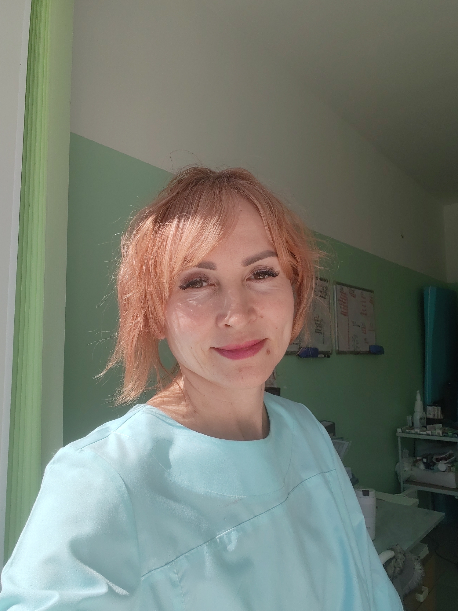 A woman with light orange hair is in a room painted light green. She has dark eyelashes and lipstick on. She wears a light blue shirt. There are white vertical blinds to her left and, behind her to the right, two whiteboards on the wall. The image may have been taken in Chaikovsky, Russia.