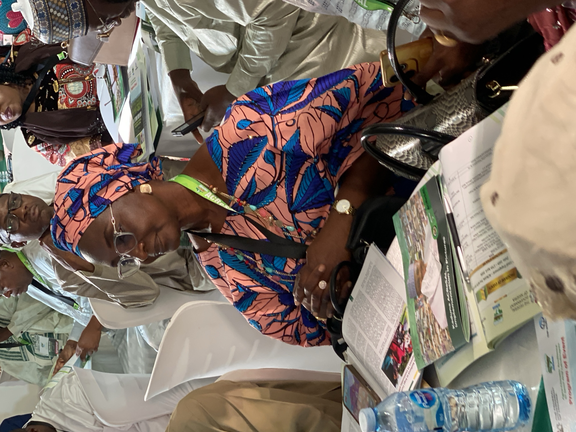 The image shows an indoor scene with several people, likely at an event or conference. One woman is in focus; she wears a colorful patterned top and headwrap, eyeglasses, a wristwatch, and rings. She is looking down at papers. There is a black snakeskin-patterned handbag on the table next to her. There is a water bottle and multiple documents with the word "Kaduna" visible on some of them. Given the presence of "Kaduna" on the documents, it is likely that the location is somewhere in the Nigerian city of Kaduna. The people are dressed in formal attire with lanyards around their necks.