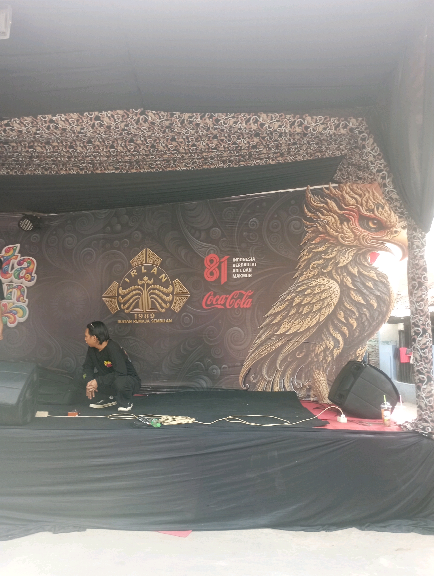 A low-angle, medium shot captures a stage set up outdoors. The stage backdrop is dark and features a large, ornate graphic of a golden eagle with intricate feather details. To the left of the eagle is a graphic with stylized lettering and what appears to be a crest with the text "IRLAN 1989 IKATAN REMAJA SEMBILAN". To the right of this crest, the number "81" is prominently displayed in red, followed by the Coca-Cola logo and the words "INDONESIA BERDAULAT ADIL DAN MAKMUR".
In the foreground, a person is kneeling on the stage, facing away from the camera and looking towards the right. They are wearing a black long-sleeved shirt and dark pants, with black sneakers. Their hands are clasped in front of them. To the left of the kneeling person is a black speaker. Another speaker is visible on the right side of the stage. Electrical cords and a power strip are on the stage floor, along with a glass with a straw and a drink.
The stage is covered with black fabric. Above the backdrop, decorative fabric with a repeating pattern is draped, and black fabric forms a canopy over the stage. The overall scene suggests an outdoor event or performance. The image's composition and style do not provide specific indicators to pinpoint the exact location within East Jakarta, Indonesia, beyond it being an outdoor public space likely hosting an event.
