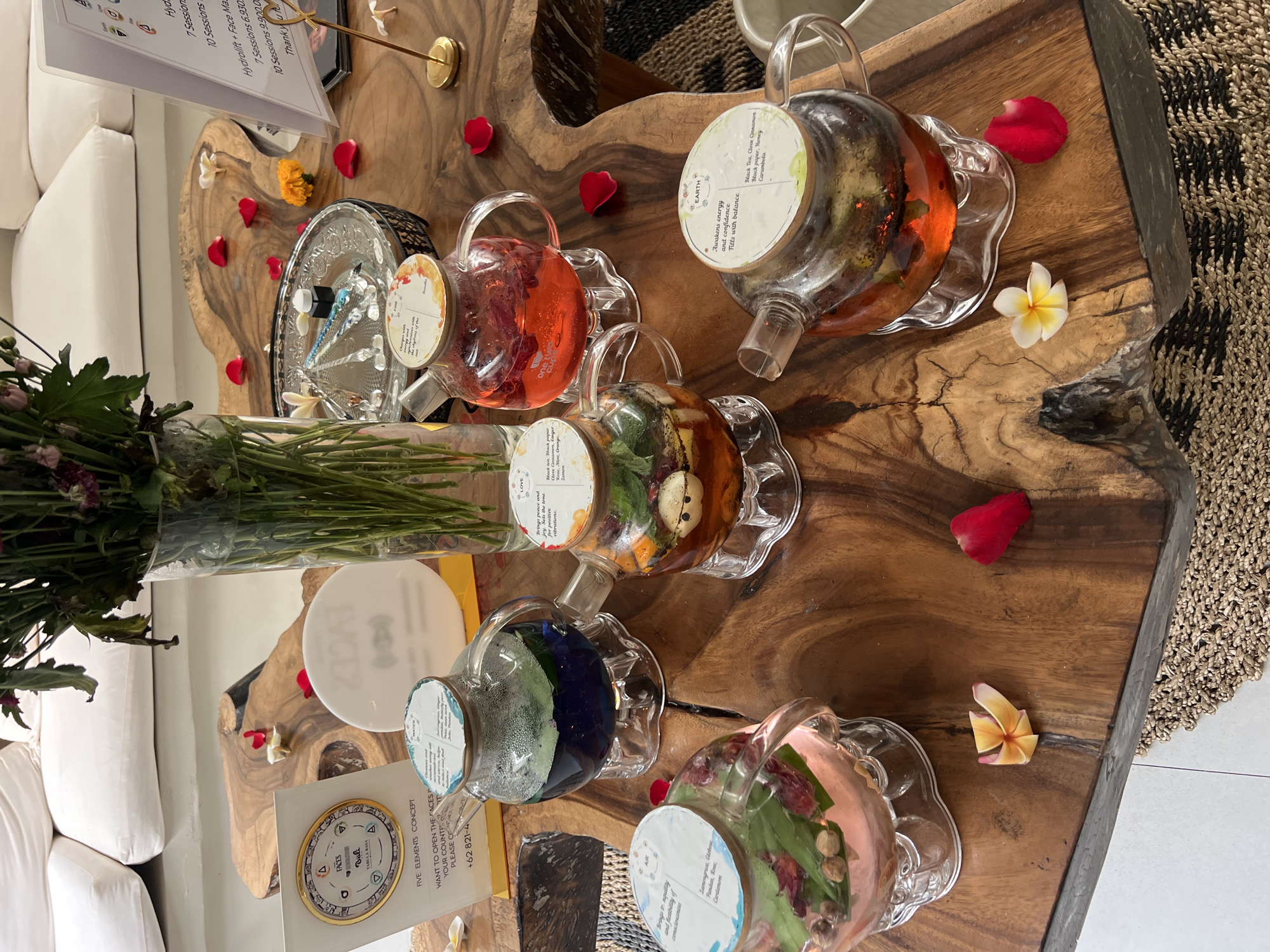 An eye-level shot captures a vibrant display of six glass teapots, each filled with different colorful infusions, arranged on a rustic, live-edge wooden table. The teapots contain various fruits, herbs, and spices, with liquids ranging from red, orange, and blue to brownish and pink. Each teapot has a circular label on its lid, indicating a theme or element.
Visible labels include "EARTH," which "Awakens energy and confidence," and "LOVE," which "Brings peace and positive vibrations." The "EARTH" infusion lists "Black Tea, Clove Cinnamon, Black pepper, Honey, Carambola." Another label, for a blue infusion, mentions "Brings tranquility and flexibility of consciousness," with ingredients like "Lamongrass, Globosc, Pandan, Rose, Cardamom," likely representing "AIR."
The table is adorned with red rose petals, delicate white and yellow plumeria flowers, and a yellow marigold flower, adding a serene and decorative touch. A tall glass vase holding green foliage and purple flowers stands to the left. In the background, part of a white sofa or cushion is visible, alongside a woven rug, suggesting a comfortable, luxurious interior setting.
Two signs provide additional context. An upright white sign lists services and prices, such as "Hydrolift - Face Ma," with "7 Sessions 6.930" and "10 Sessions 9.900.0," indicating a spa or wellness center. A round white sign on the table reads "FACES," "Bali," "TABELA RASA," and "FIVE ELEMENTS CONCEPT," with a call to action: "WANT TO OPEN THE FACES YOUR COUNT PLEASE CO +62 821-4". The overall scene evokes a holistic wellness experience, likely in Bali, focusing on natural elements and therapeutic infusions.