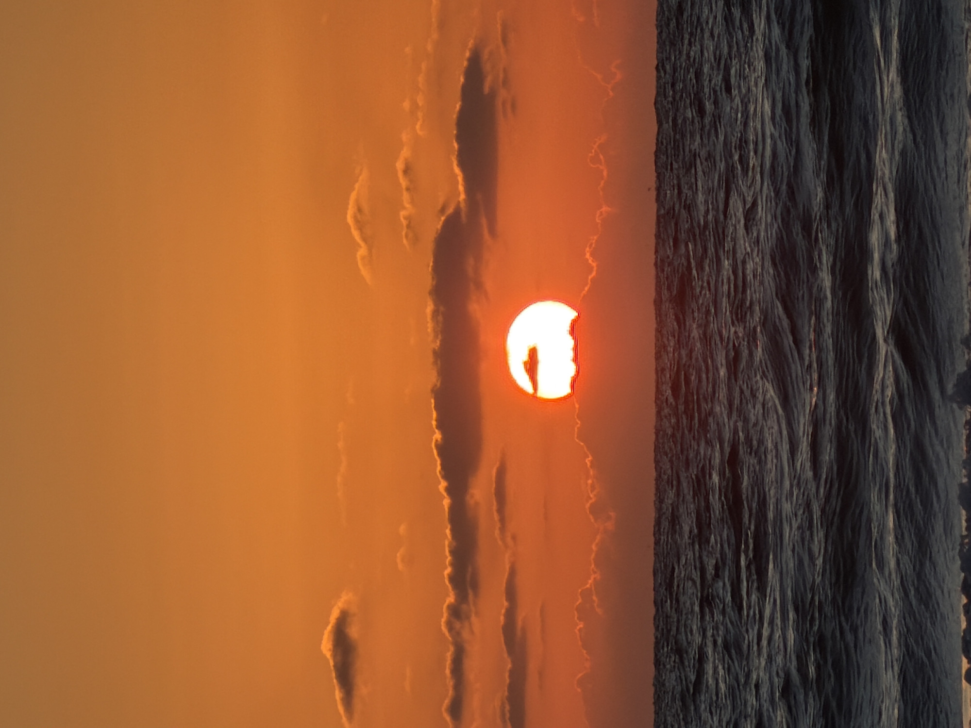 The image shows a sunset in Tel-Aviv, Israel. The sky is orange, with dark clouds and the sun. A dark textured vertical barrier occupies the right side of the frame. The horizon is unclear.