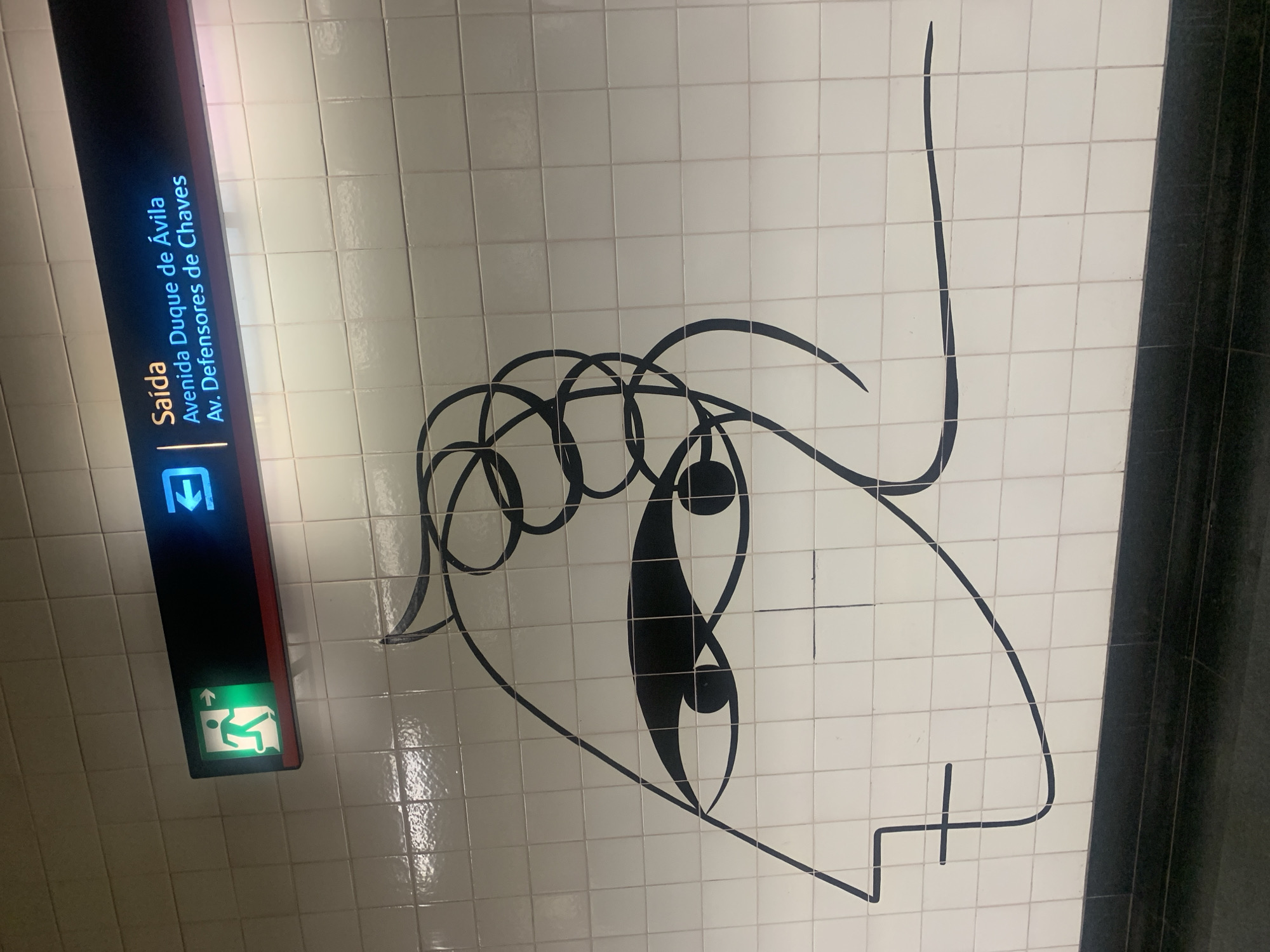 The image shows a white tiled wall in a Lisbon Metro station, specifically at the Saldanha station. The wall features a minimalist black line drawing of a face. Above the drawing, a black sign with illuminated text indicates "Saída" (Exit) in Portuguese, with arrows pointing left. The text below "Saída" reads "Avenida Duque de Ávila" and "Av. Defensores de Chaves," indicating directions from the station. To the left of the text, a green illuminated exit sign with a running figure is visible. The floor in the foreground is dark and appears to be a platform.