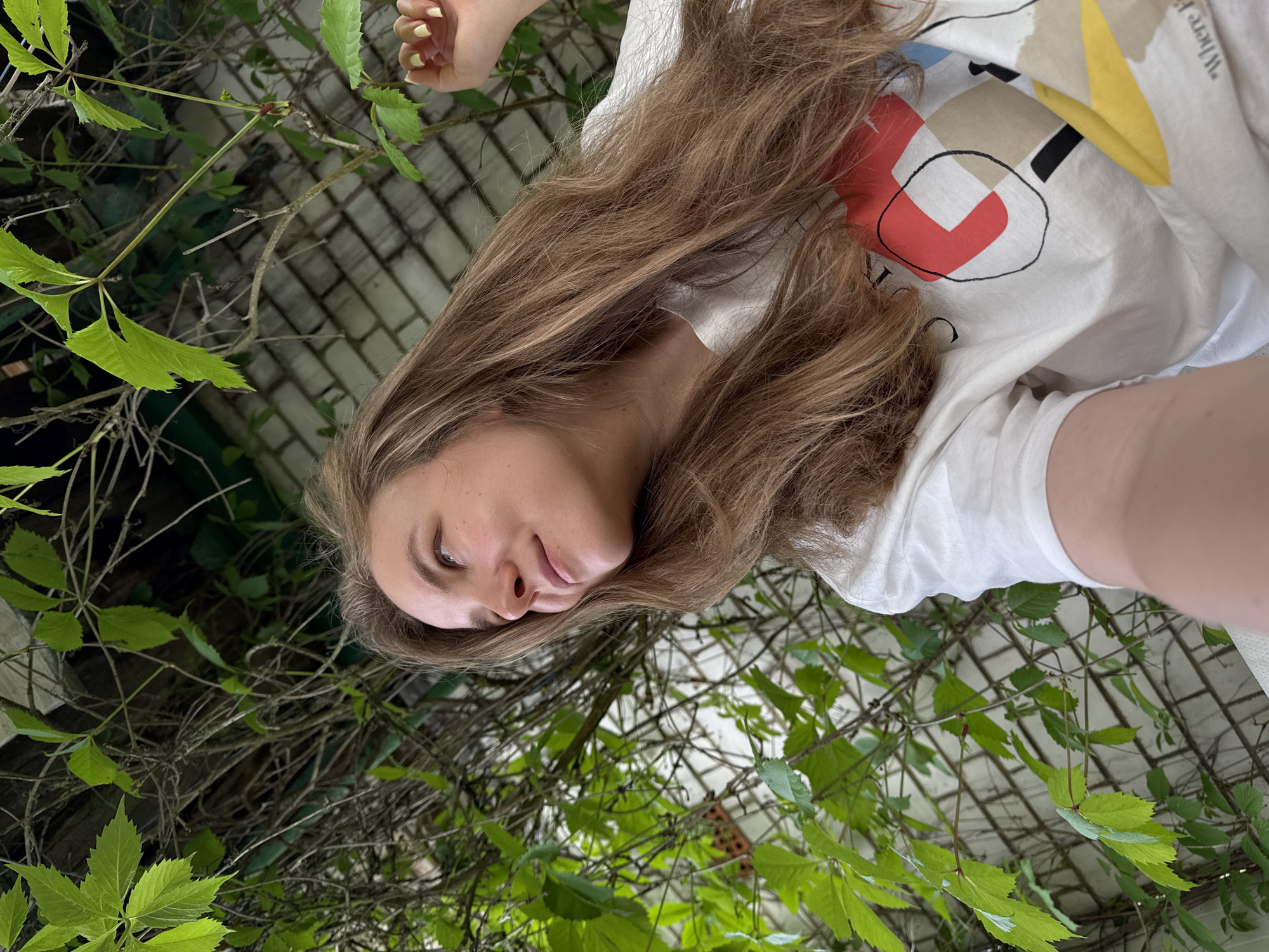 A young woman with long, wavy light brown hair is depicted in a bright outdoor setting, her face in soft focus. She is looking slightly upwards and to the side with a calm, serene expression and a subtle smile. She is wearing a white t-shirt featuring an abstract design with bold red, black, yellow, and beige geometric shapes. Part of a text, "Where I", is visible on her shirt. Her left hand, with light yellow nail polish, is visible in the upper left corner of the frame, suggesting she might be taking a selfie.
The background is filled with abundant green foliage, appearing to be climbing vines or creepers (like Virginia Creeper), which partially obscure a light-colored brick wall and a white lattice structure. The natural, bright lighting indicates it is daytime. The overall mood is peaceful and natural. The location is specified as Красный Маяк, Russia.