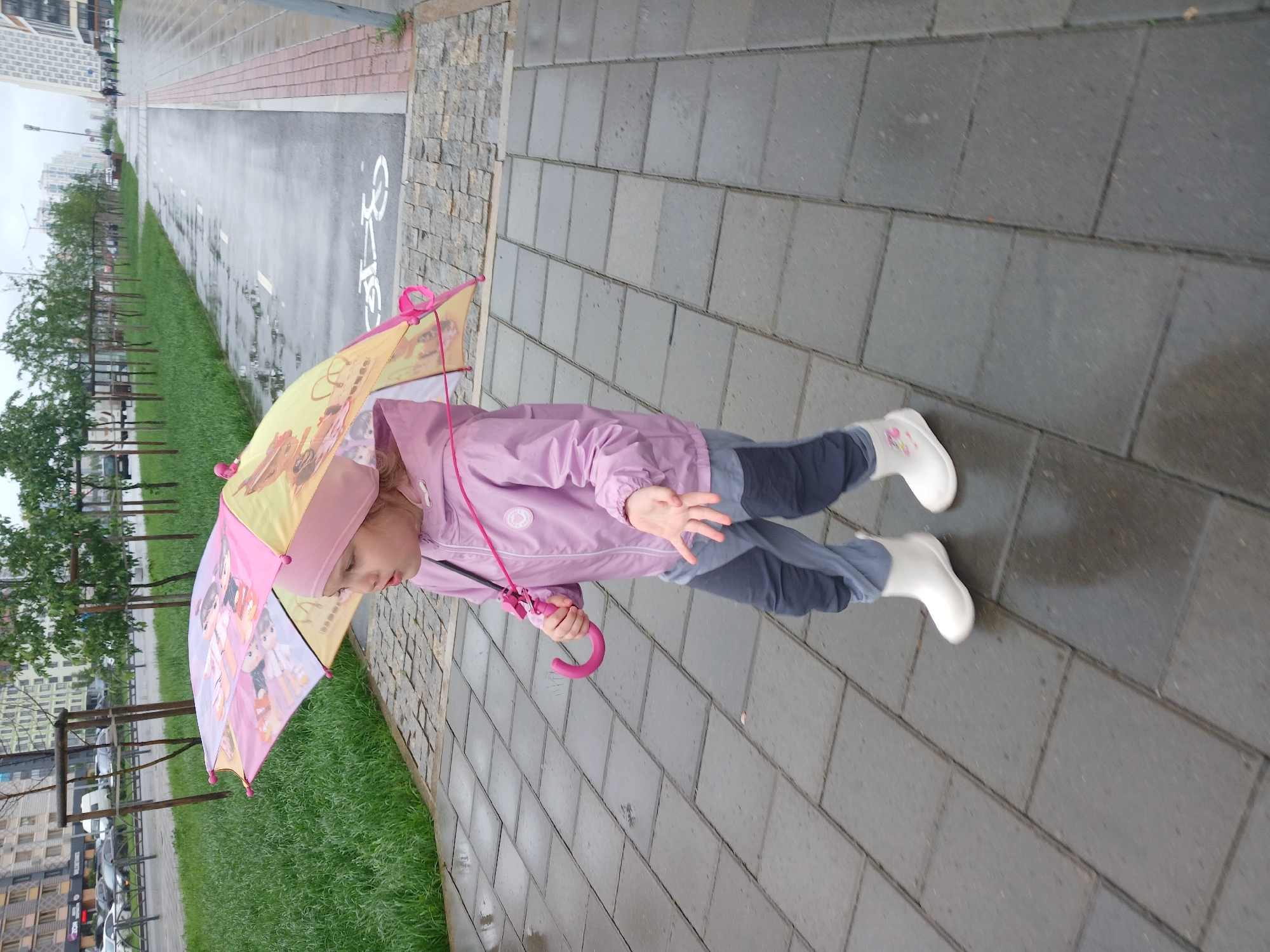 A young girl in a pink jacket, grey pants, and white boots walks on a wet, gray-tiled sidewalk, holding an open, colorful umbrella featuring cartoon characters. The sidewalk is wet, indicating recent rain. To the left of the sidewalk, there is a strip of green grass, and beyond that, a street with parked cars and apartment buildings. To the right of the sidewalk, there is a wet asphalt path with white markings, including a bicycle symbol, and further on, more apartment buildings. The sky is overcast. This scene is consistent with a residential area in Yekaterinburg, Russia, during inclement weather.