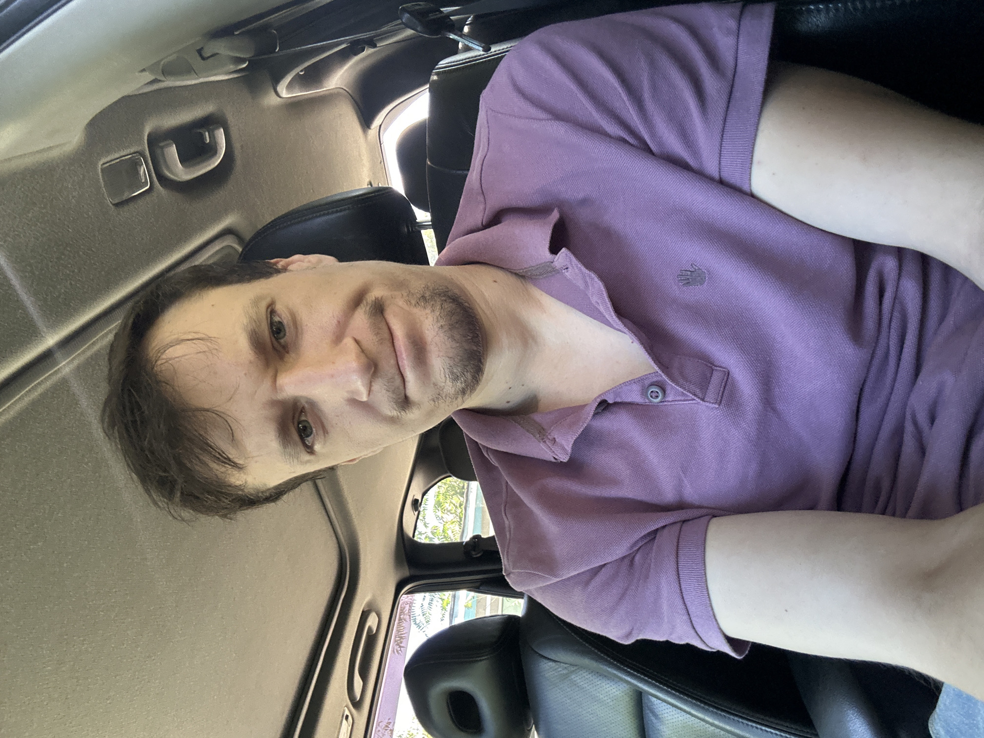 A man sits in the driver's seat of a car with a sunroof open, looking directly at the camera. He has brown hair and a goatee, and is wearing a purple polo shirt. The interior of the car is visible, including black leather seats and the car's headliner. Outside the car, trees and foliage are visible through the windows, suggesting an outdoor setting. The specific location in Makiv, Ukraine, cannot be determined from the image alone.