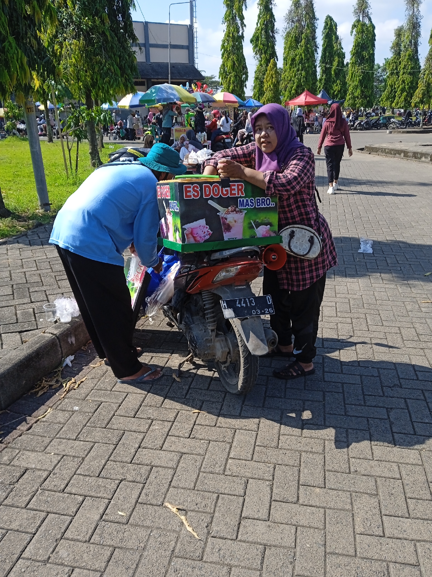 The scene is a street market in Kebondalem, Indonesia. A man in a light blue shirt and a blue bucket hat is preparing "Es Doger" (an Indonesian shaved ice dessert) from a box attached to the back of a parked orange Honda motorcycle with license plate H 4413 D expiring in March 2026. A woman wearing a purple hijab and a pink plaid shirt leans against the "Es Doger" box, seemingly the vendor or assistant. The market stretches into the background with various vendor stalls under colorful umbrellas and numerous parked motorcycles. People are walking around the market, including one woman in a maroon shirt and black pants. Tall green trees line the edge of the parking lot. The ground is paved with gray rectangular bricks. Litter, including plastic cups and bags, is visible on the ground.