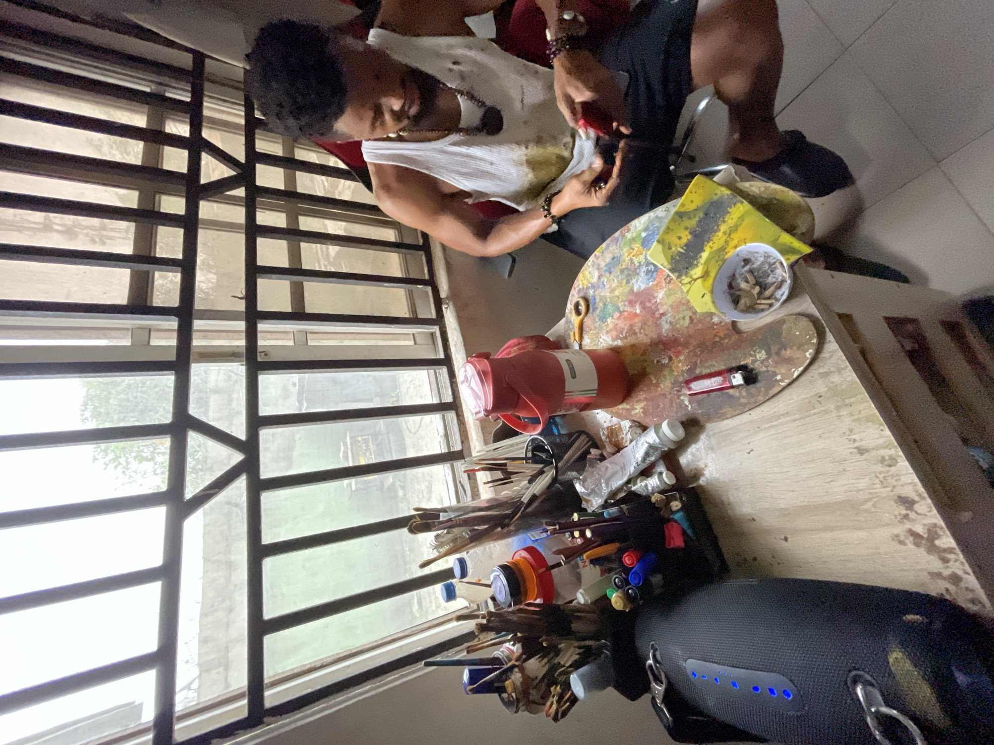 The image depicts an indoor scene, possibly an artist's studio, in Ado, Nigeria. A young Black man, wearing a white tank top and black shorts, is seated at a wooden desk. His tank top has stains, suggesting he is engaged in artistic work. He is holding a red object in his hands, focused on his task. The desk is cluttered with art supplies, including brushes, paints, tubes of what appears to be toothpaste or paint, markers, and bottles. A red thermos and a wooden palette splattered with various colors of paint are also prominent on the desk. To the left of the desk, a large black speaker with blue LED lights is visible. Behind the desk, a barred window offers a view of the outside, with glimpses of a concrete wall and greenery. The overall environment suggests a working space dedicated to creative pursuits.