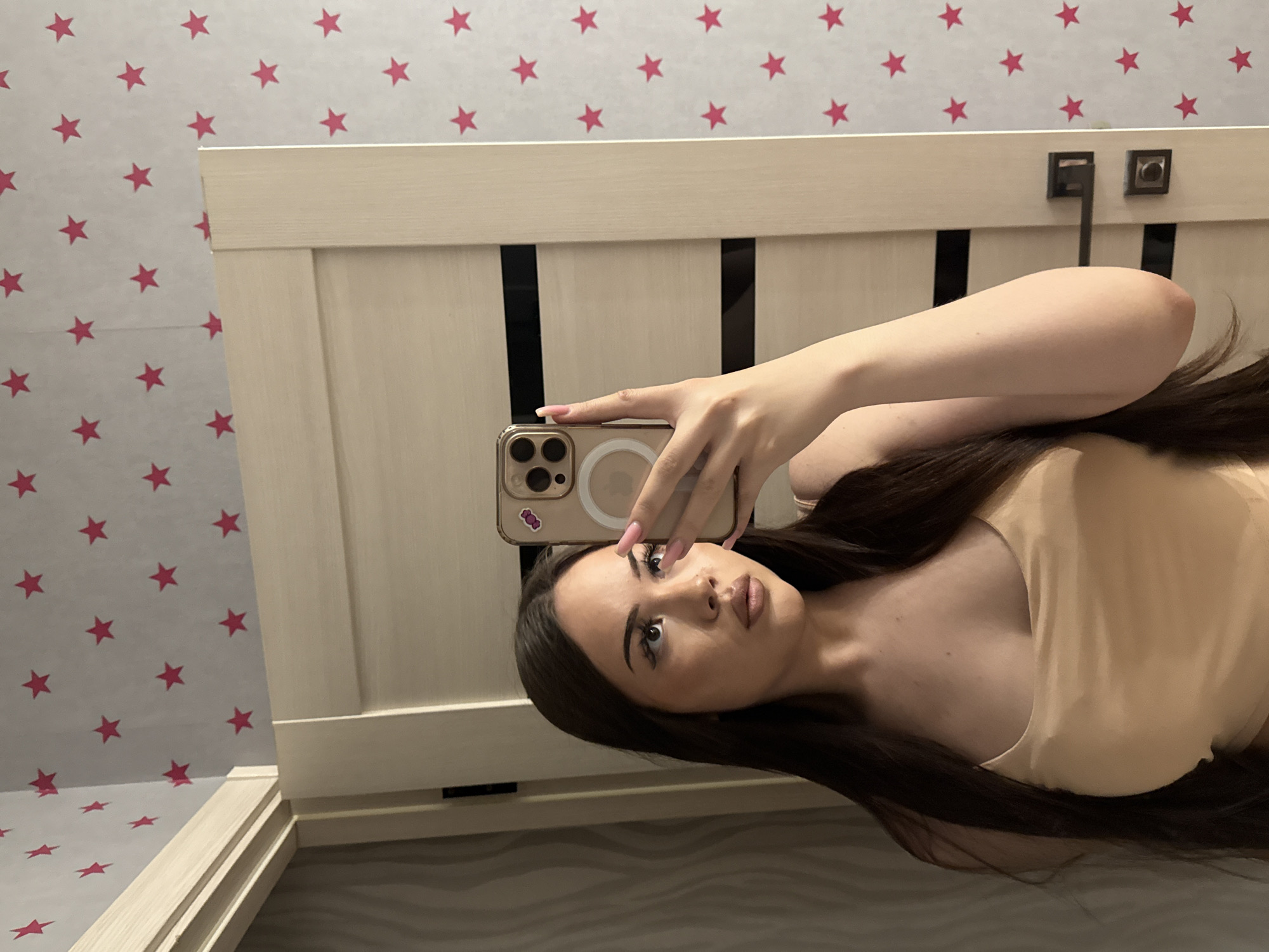 A young woman is captured in a mirror selfie, with the image rotated 90 degrees clockwise. She has long, dark brown hair and is wearing a light, possibly beige or peach-colored, sleeveless top. Her face is made up with eyeliner and lipstick, and she gazes directly into the camera of her smartphone. She holds a modern smartphone, identifiable as an iPhone by its camera configuration, in a clear case with a visible MagSafe ring. A small purple-pink graphic sticker adorns the back of the phone case. Her fingernails are manicured with a light pink polish.
The scene is an indoor setting, likely a bedroom in Naberezhnye Chelny, Russia. The background features distinctive wallpaper with a repeating pattern of red stars against a light gray or white backdrop. Below the wallpaper, a light-colored wooden headboard or bed frame is visible, which includes dark vertical accents and a metallic fixture on its upper edge. The woman's pose suggests she is lying down, taking the photo of her reflection. There is no legible text visible in the image.