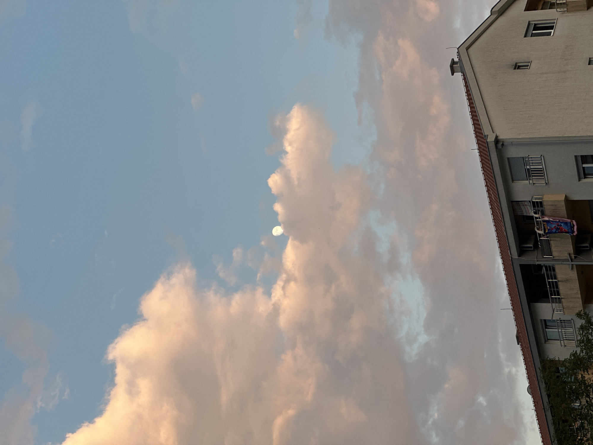 The image is a vertical, low-angle shot of a building against a twilight sky filled with large, fluffy clouds. The building, a light-colored multi-story structure, dominates the right side of the frame. It has multiple windows and balconies with metal railings. On one balcony, a colorful piece of laundry, possibly a blanket or sheet with a graphic design, is visible. The building's roofline, with red tiles and a chimney, is also prominent.
The sky is a pale blue, transitioning to soft pinks and grays, suggesting it is either early morning or late evening. The clouds are voluminous and illuminated by the setting or rising sun, giving them a warm, peachy glow in places. Towards the upper left of the frame, a small, bright orb, which appears to be the moon, is visible through a gap in the clouds. A few small birds are also faintly visible in the sky.
The scene suggests a residential setting in Budva, Montenegro, likely a city or town with apartment buildings. The overall mood is serene and tranquil, capturing a quiet moment in the transition between day and night. There is no visible text in the image.