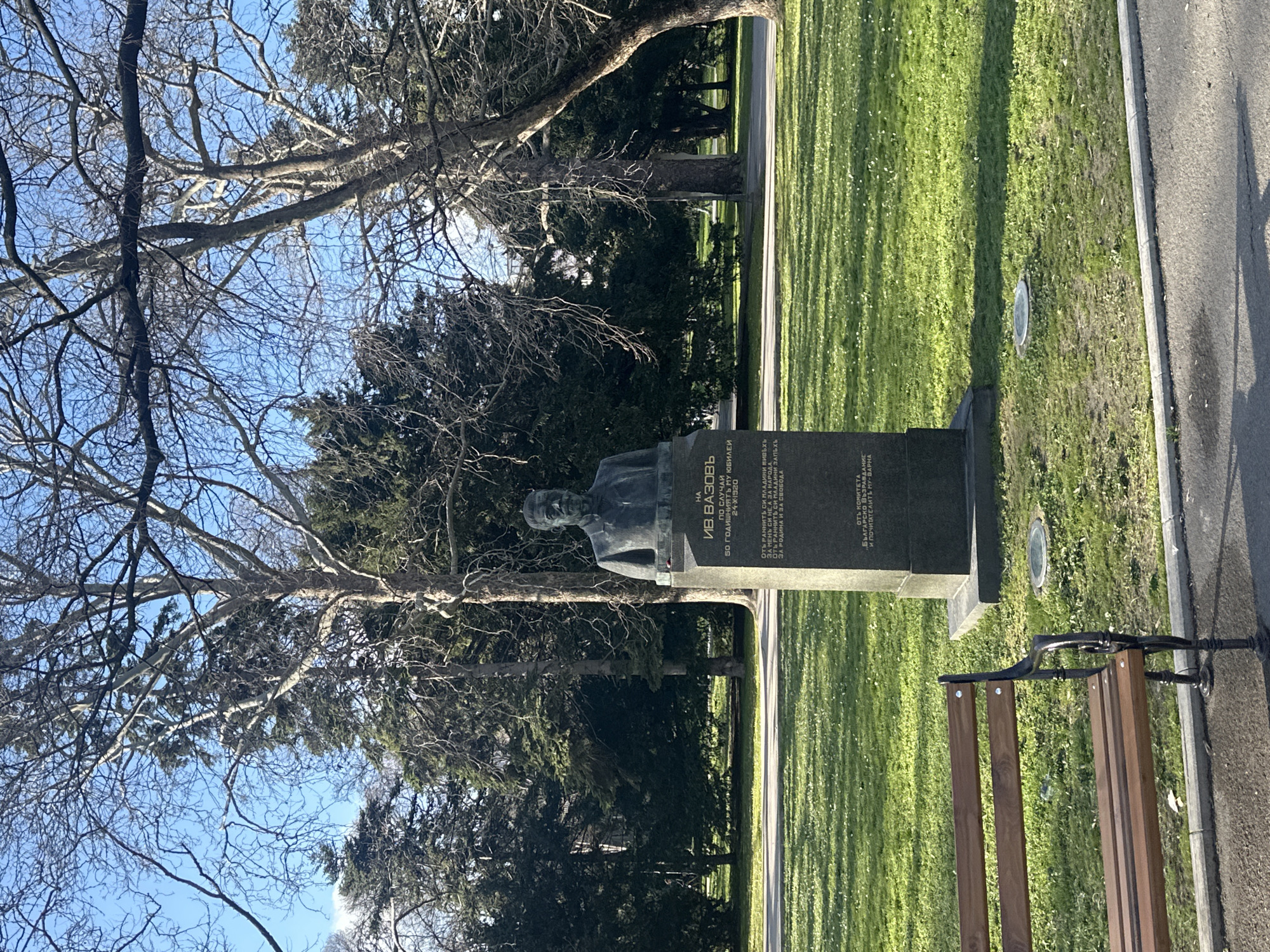 A daytime outdoor scene depicts a bronze bust of Ivan Vazov atop a dark gray stone pedestal, situated in the Sea Garden (Morska Gradina) of Varna, Bulgaria. The bust, facing forward, portrays a man with a mustache, wearing a suit jacket and tie. The pedestal's inscription in Cyrillic identifies the subject as "ИВ. ВАЗОВ" (IV. VAZOV), commemorating his 50th anniversary on October 24, 1920, and notes its dedication from "БЪЛГАРСКО ВЪЗРАЖДАНЕ И ПОЧИТАТЕЛИТЕ МУ - ВАРНА" (Bulgarian Revival and his admirers - Varna).
The monument stands on a bright green lawn, speckled with small white flowers, under clear, direct sunlight. Shadows are visible across the grass. A circular metal cover is embedded in the grass near the monument's base. In the foreground, a dark asphalt path runs horizontally, bordered by a light gray concrete curb. A wooden park bench with black, ornate metal supports is positioned on this path in the lower-left, partially cropped. The background features a dense collection of trees; tall deciduous trees with bare branches are prominent, interspersed with dark green coniferous trees. The sky above is clear and blue. No individuals are present in the frame.