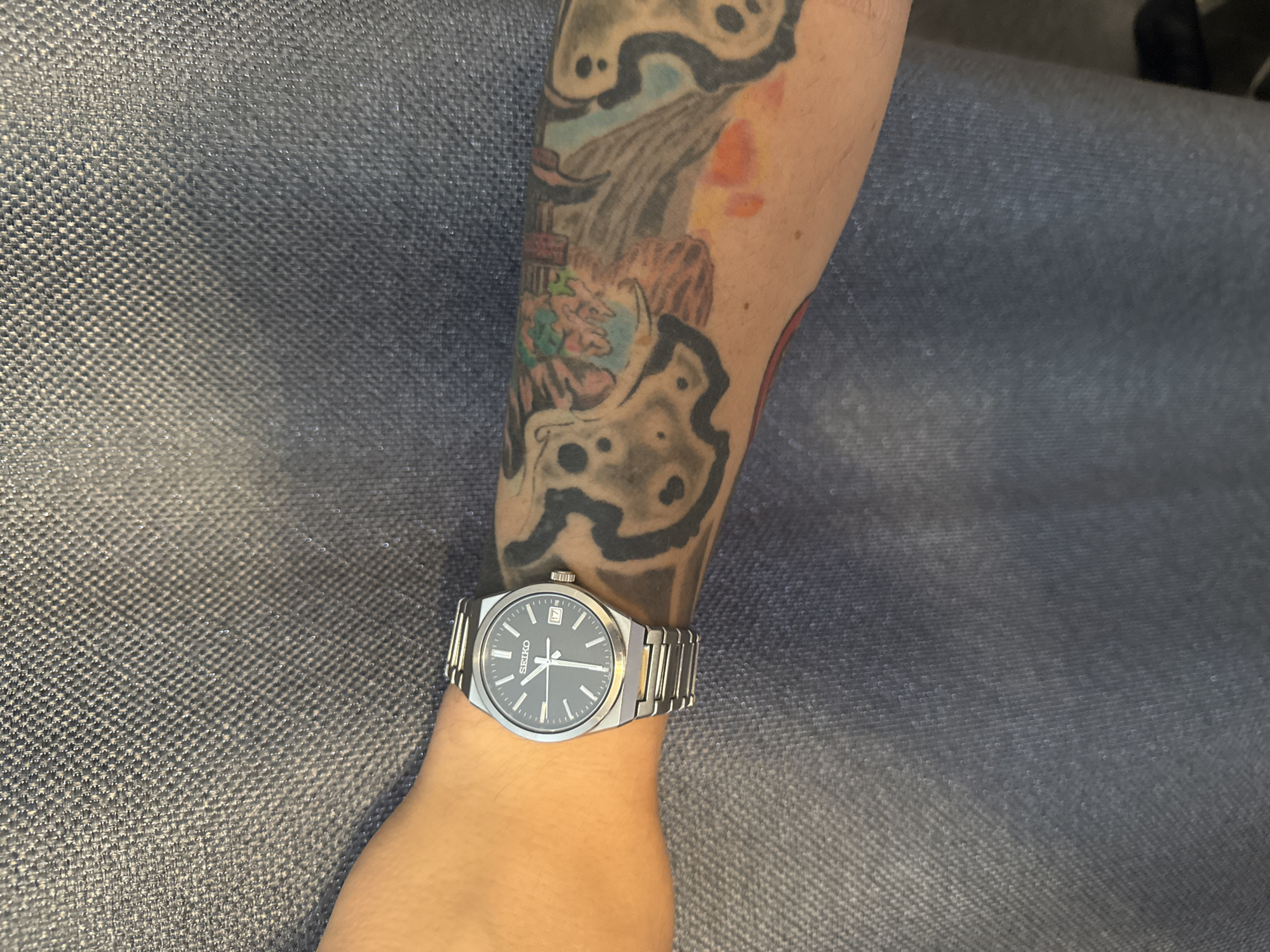 The image shows a close-up view of a person's left arm, which is adorned with a tattoo and a wristwatch. The arm rests on a textured, dark blue fabric, possibly clothing or upholstery. The wristwatch is a silver Seiko with a dark dial, featuring white hour markers and hands, and a date window displaying "17". The tattoo is a black and grey design with hints of color, extending along the forearm. The background is a blurry, dark blue textured surface. The lighting suggests an indoor environment. The location in Vladivostok, Russia is not discernible from the visual information provided.