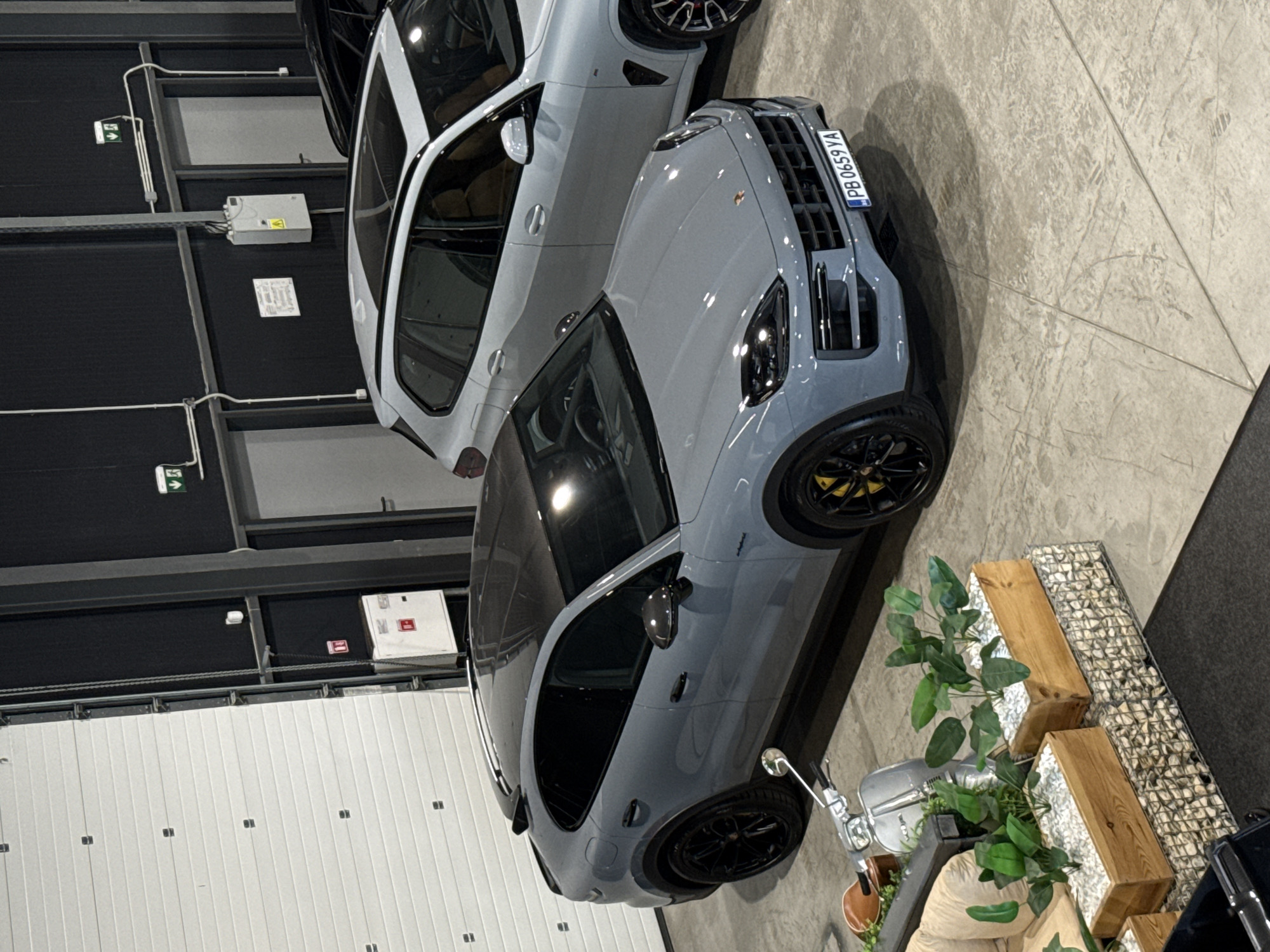 The image depicts an indoor showroom or garage in Plovdiv, Bulgaria. The focal point is a grey Porsche Cayenne Coupe parked on a concrete floor. To its right, a light-colored SUV, possibly a BMW X5, is partially visible. In the background, a large white garage door occupies the left side of the frame, with industrial shelving and lighting fixtures on the wall. A silver Vespa scooter is parked in front of the Porsche, accompanied by two wooden planters filled with greenery placed on gabion retaining walls. The environment appears to be well-lit, likely with artificial lighting, creating reflections on the car's surface. The overall scene suggests a car dealership or a private collection space.