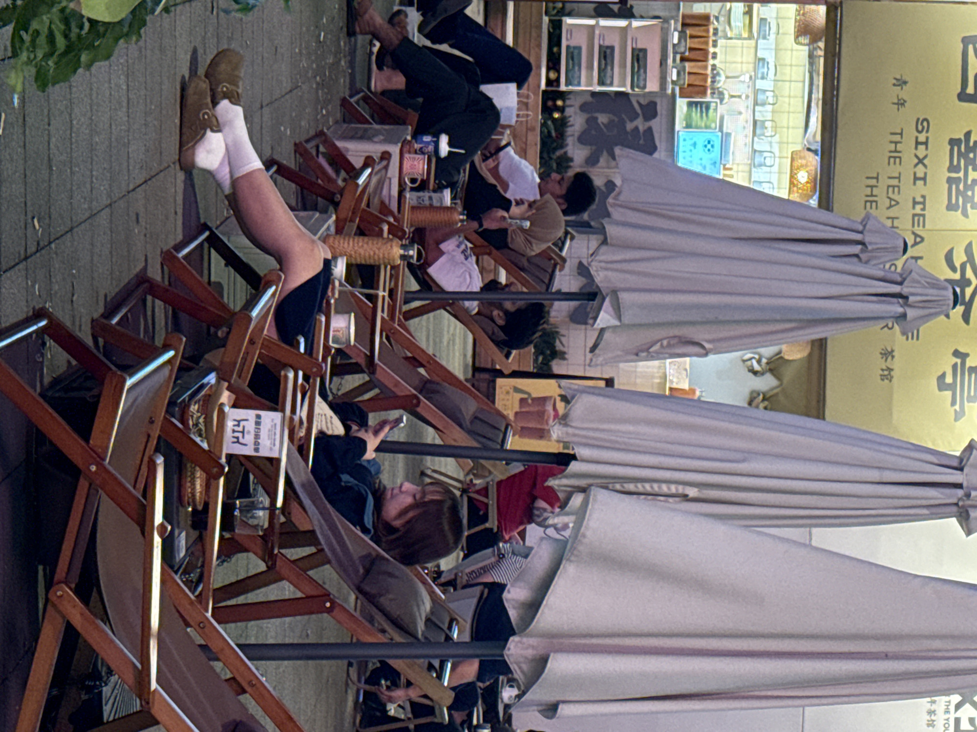 The image shows people lounging in wooden chairs outdoors, with their legs propped up, possibly at an outdoor café or lounge. Several people are seated, some using their phones. White umbrellas provide shade for the patrons. The location appears to be in the Yuzhong District of China, as indicated by the Chinese text on a nearby sign that reads "SIXI TEA H" along with other Chinese characters. The sign also includes the English phrase, "THE TEA H". Additional details visible are the wooden chairs, tables with drinks and other objects, and a shop front in the background.