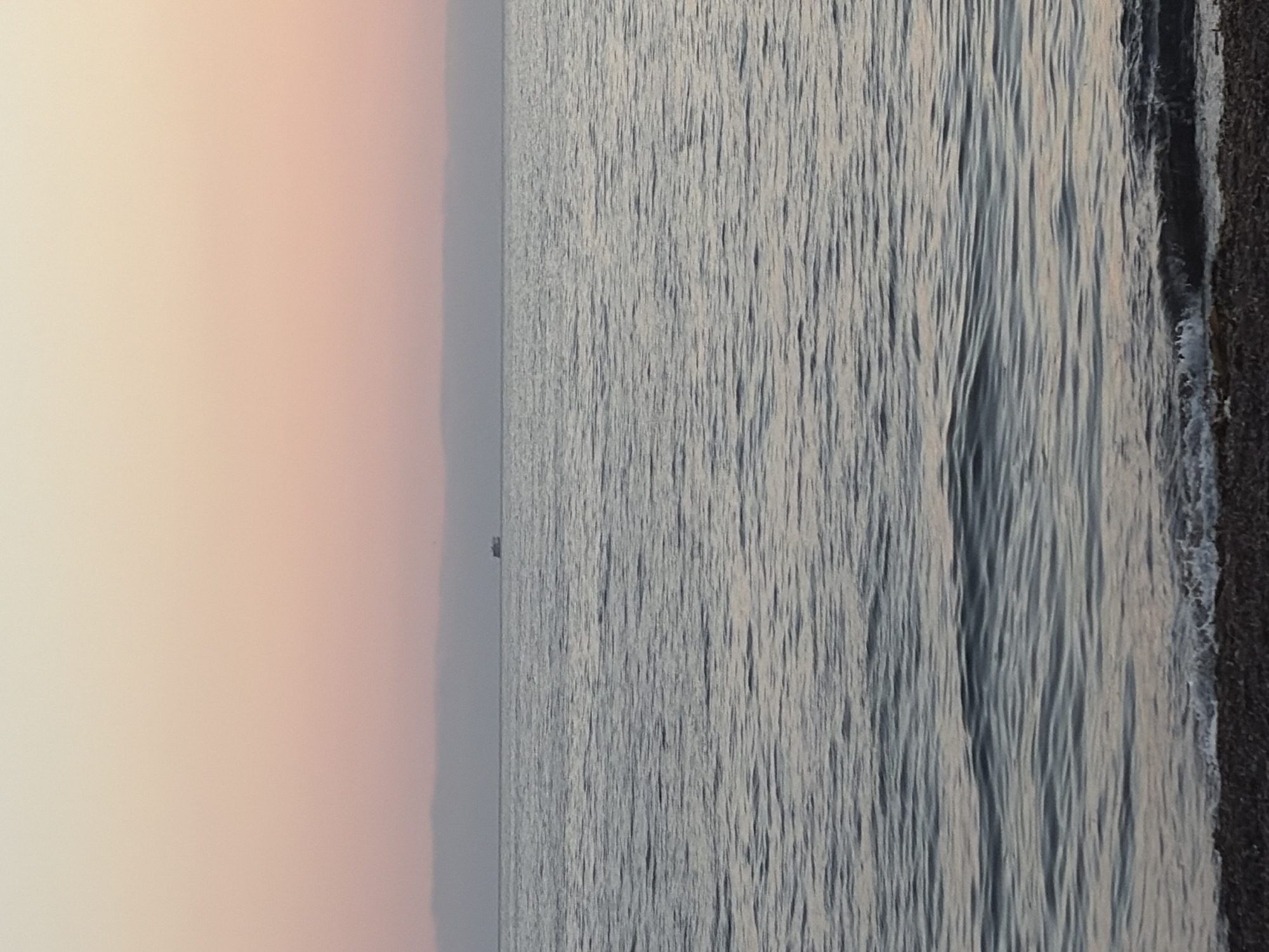This is a photograph of a calm sea at dusk. The sky is a gradient of pale yellow at the top transitioning to soft pink and then a muted purple near the horizon. A thin band of hazy clouds is visible on the horizon. In the distance, a small, dark-colored boat is visible on the water. The sea itself is a calm, rippling expanse of gray-blue water, reflecting the soft light of the sky. In the foreground, a narrow strip of dark pebble beach is visible, with small waves gently lapping at the shore. The overall impression is one of tranquility and the end of a day. The image does not depict any people or specific identifiable landmarks in Ozerskoe, Russia, but captures a general coastal scene consistent with such a location.