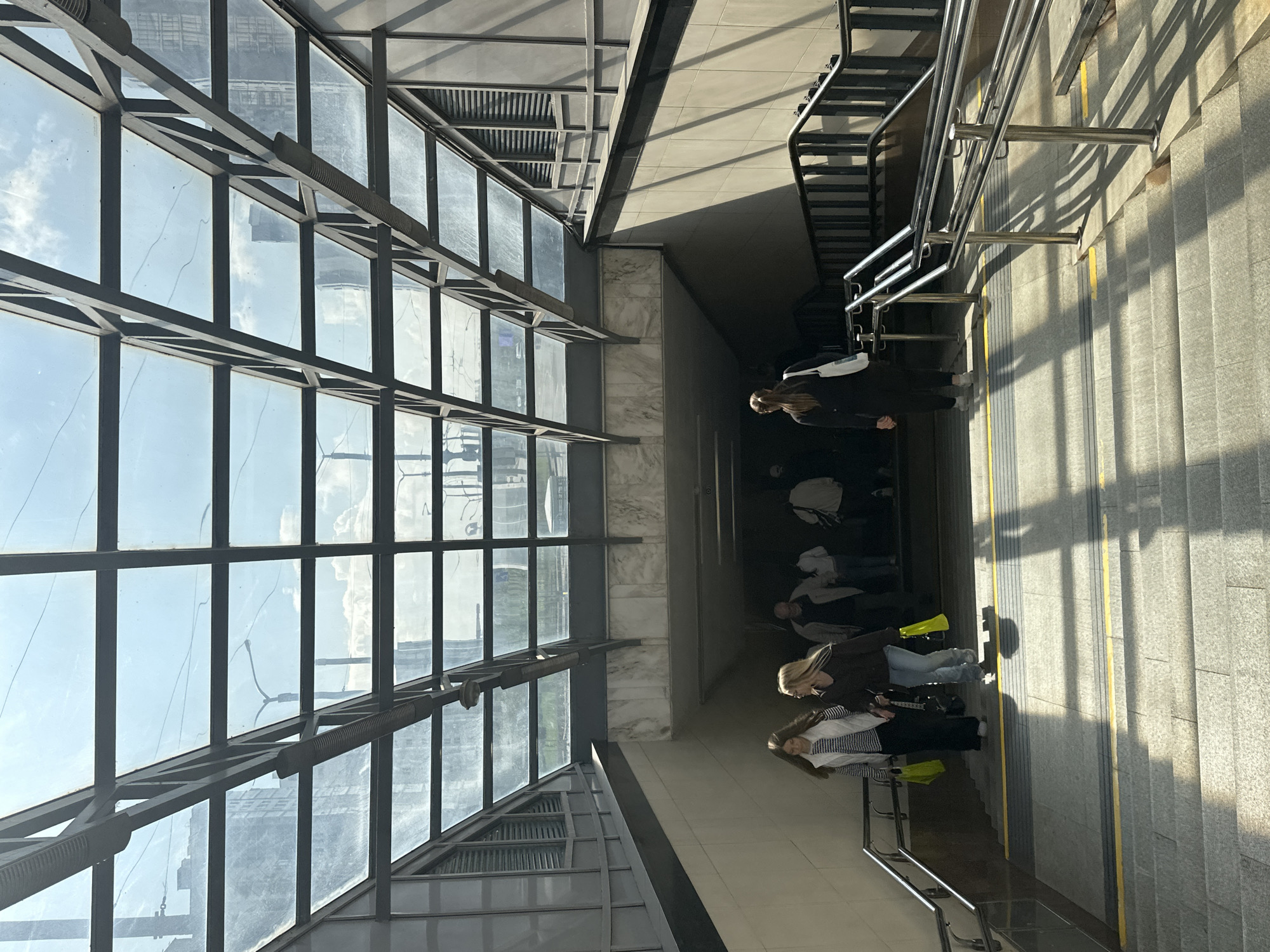The image depicts the entrance to an underpass or subway station in Minsk, Belarus. The structure features a modern glass and metal roof, allowing natural light to illuminate the wide, concrete staircase leading down into the passage. Several individuals are visible, descending the stairs. On the left, two women walk side-by-side, one wearing a white vest over a striped shirt and the other in a dark top, both carrying yellow shopping bags. Further down the stairs, a lone woman in black with a white tote bag is heading into the underpass, while other figures are indistinctly visible in the background, suggesting pedestrian traffic. The walls of the underpass are lined with light-colored tiling, and metal railings guide the descent. Sunlight casts strong shadows across the stairs, creating a dramatic play of light and shade. The overall environment suggests a public transportation hub in an urban setting.