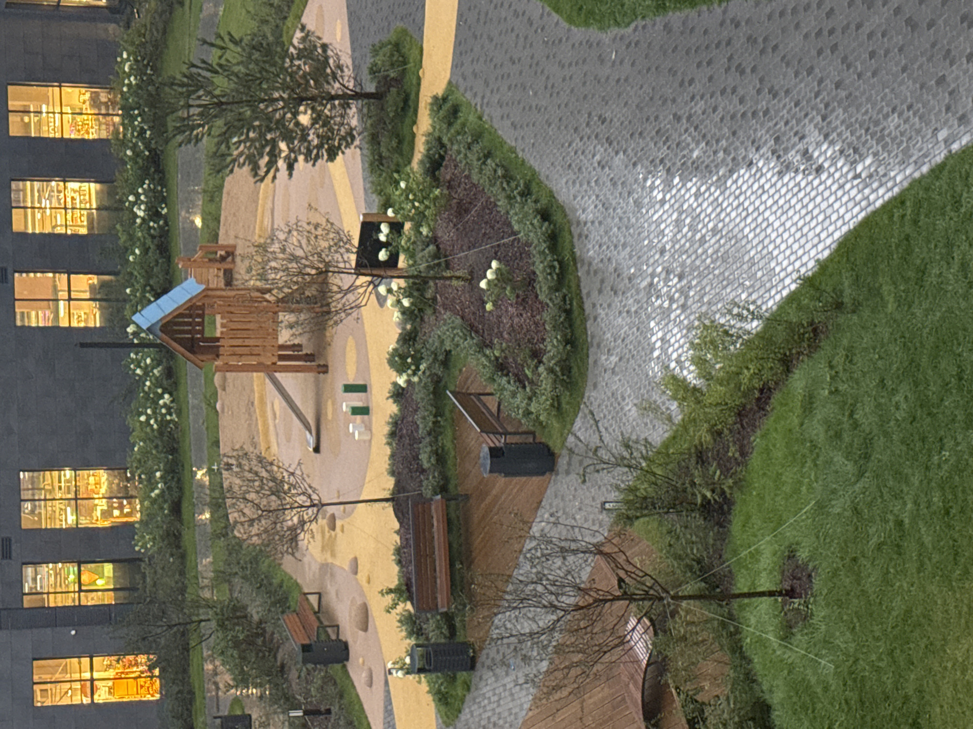 This image captures an aerial view of a modern playground and courtyard in the city of Kommunarka, Russia. The scene is characterized by a combination of natural elements and urban infrastructure. A wooden playground structure with a blue roof and a slide is centrally located in a sand-filled area, surrounded by yellow and brown paving. Nearby, two park benches and trash receptacles are situated on paved walkways. Green grass, trees, and flowering bushes are interspersed throughout the area, with a dark-colored building featuring illuminated windows forming the backdrop. The ground, particularly the paved sections, appears wet, suggesting recent rainfall. The overall impression is one of a well-maintained, contemporary outdoor public space designed for recreation and relaxation.