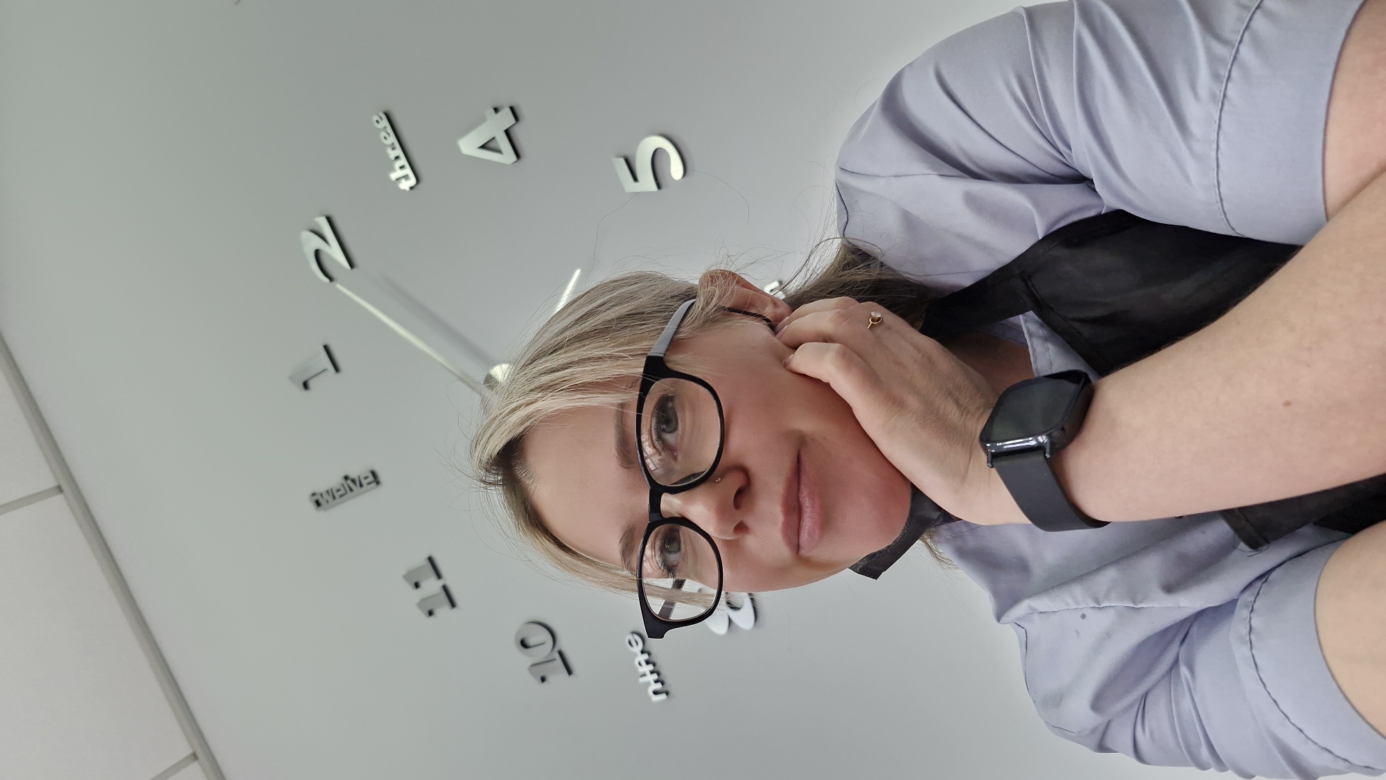 A woman with blonde hair and glasses is posing for a selfie in front of a wall clock. The clock has large, individual numerals in silver and is set at approximately 12:08. The woman is wearing a light blue shirt, a black apron, and a black smartwatch on her left wrist. Her left hand is raised near her ear. The location of the image is unknown and there is no indication that this is in Otchor, Russia.