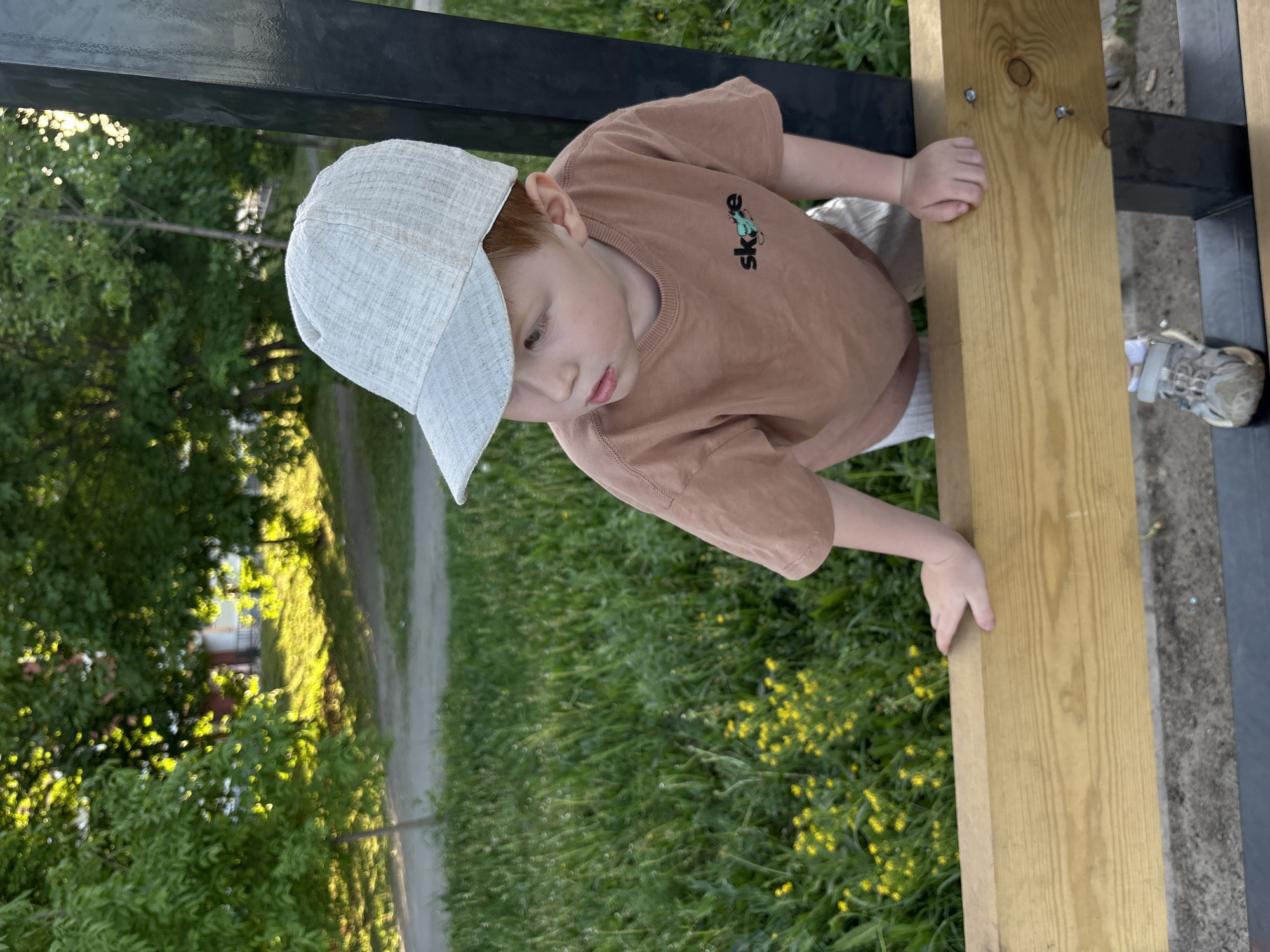 A young boy, wearing a light-colored baseball cap and a brown t-shirt with the word "skate" on it, is standing next to a wooden structure supported by a black metal frame. He is holding onto the wooden plank with his hands. Grass with small yellow flowers is visible in the background. A house is visible behind the grass, through the trees. There is a path to the left of the boy. The location may be in the city of Izhevsk, Russia.