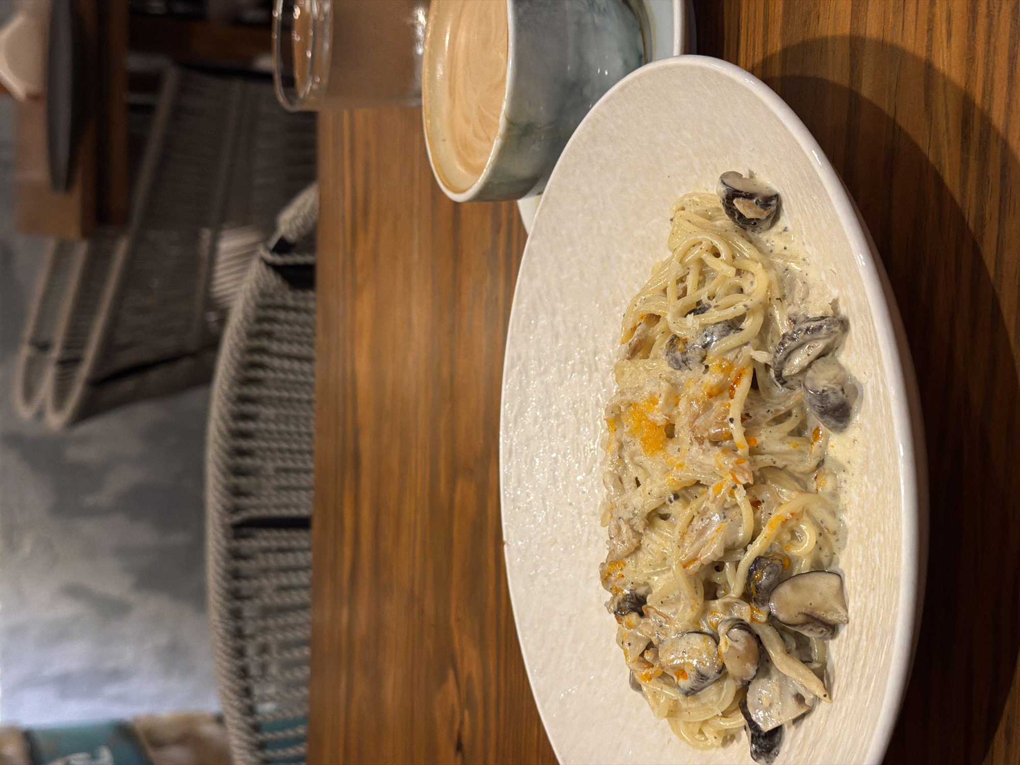 On a wooden table, a white plate holds spaghetti with mushrooms in a creamy sauce, along with a cup of coffee or latte. The scene is inside what appears to be a café or restaurant, possibly in Nha Trang Ward, Vietnam, based on the provided information. A chair is partially visible in the background.