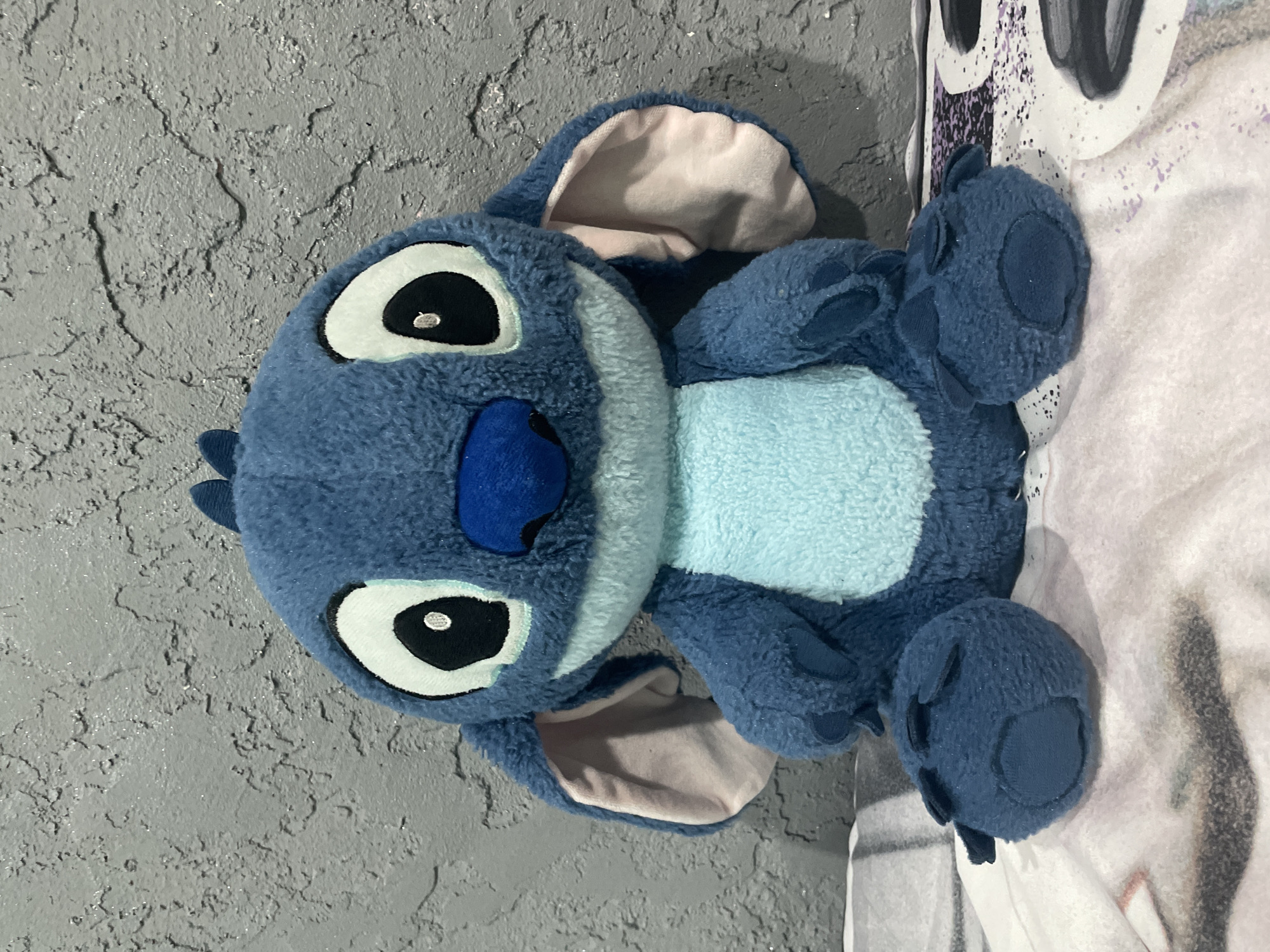 A plush blue "Stitch" toy is on a bed next to a gray wall. The location in Sofia, Bulgaria, is not discernible from the image.