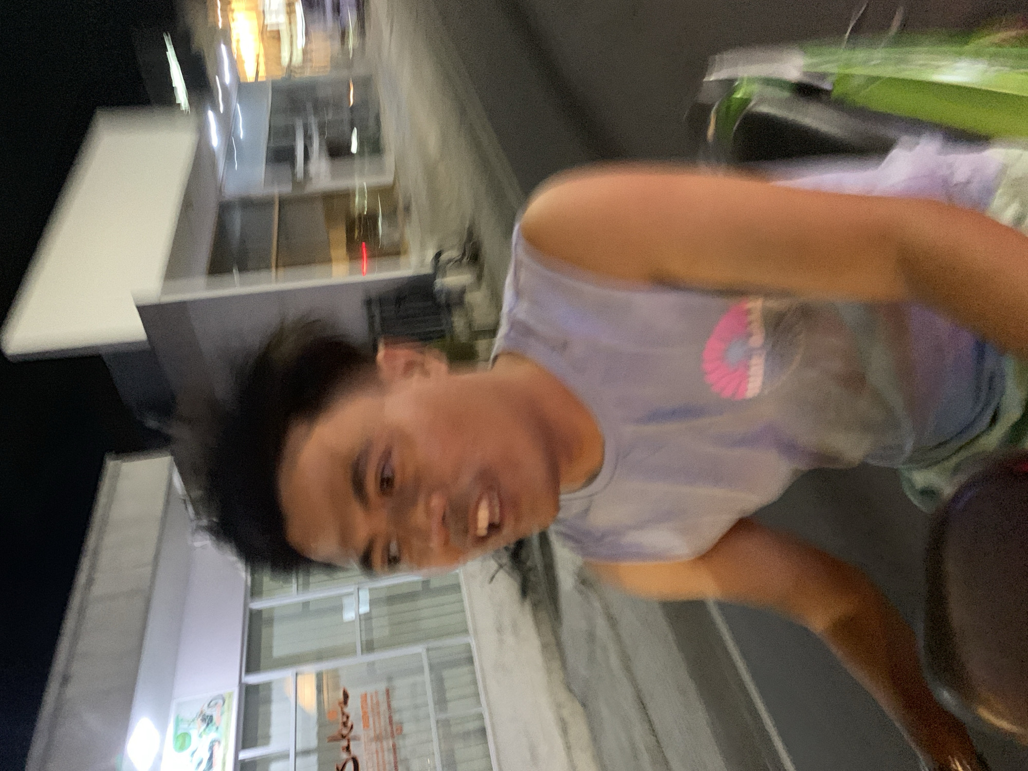 A blurred photo depicts a person wearing a light blue tank top outdoors at night. The person faces the camera with a slight grimace. A building with a lighted sign reading "Sakura" is visible in the background. The location is possibly in North Kuta, Indonesia, as suggested by the sign's language or style. Other buildings are visible in the background, illuminated by street lights. The picture appears to have been taken while in motion.