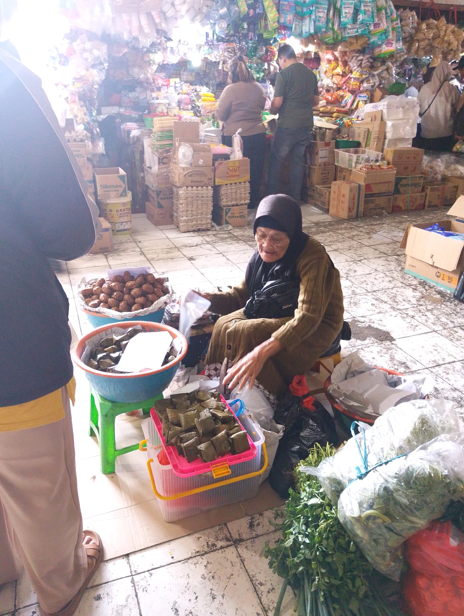 The image depicts a bustling market scene, likely in Lembang, Indonesia, with an elderly woman at the center of the activity. She is seated on a low stool, wearing a dark headscarf and a long, earth-toned garment, and appears to be engaged in selling goods. Her hands are visible, resting on items in front of her, suggesting interaction with customers or preparation of her wares.
Several baskets and containers are placed around her. One large blue-green basin holds numerous small, brown, spherical items, possibly nuts or snacks. Another, similar blue basin contains rectangular, wrapped food items, each placed neatly on white paper. A bright pink and yellow plastic container is also filled with these wrapped items. Nearby, large bundles of green leafy vegetables are visible, some wrapped in plastic.
The background reveals a crowded market environment. Stalls are laden with various products, including packaged goods hanging from above, stacks of boxes, and containers of different sizes and colors. The floor is tiled and appears somewhat worn. Other individuals are present in the scene, though their interactions are not clearly defined; some are standing and looking, contributing to the sense of a lively marketplace. The overall impression is one of everyday commerce in a local Indonesian setting.