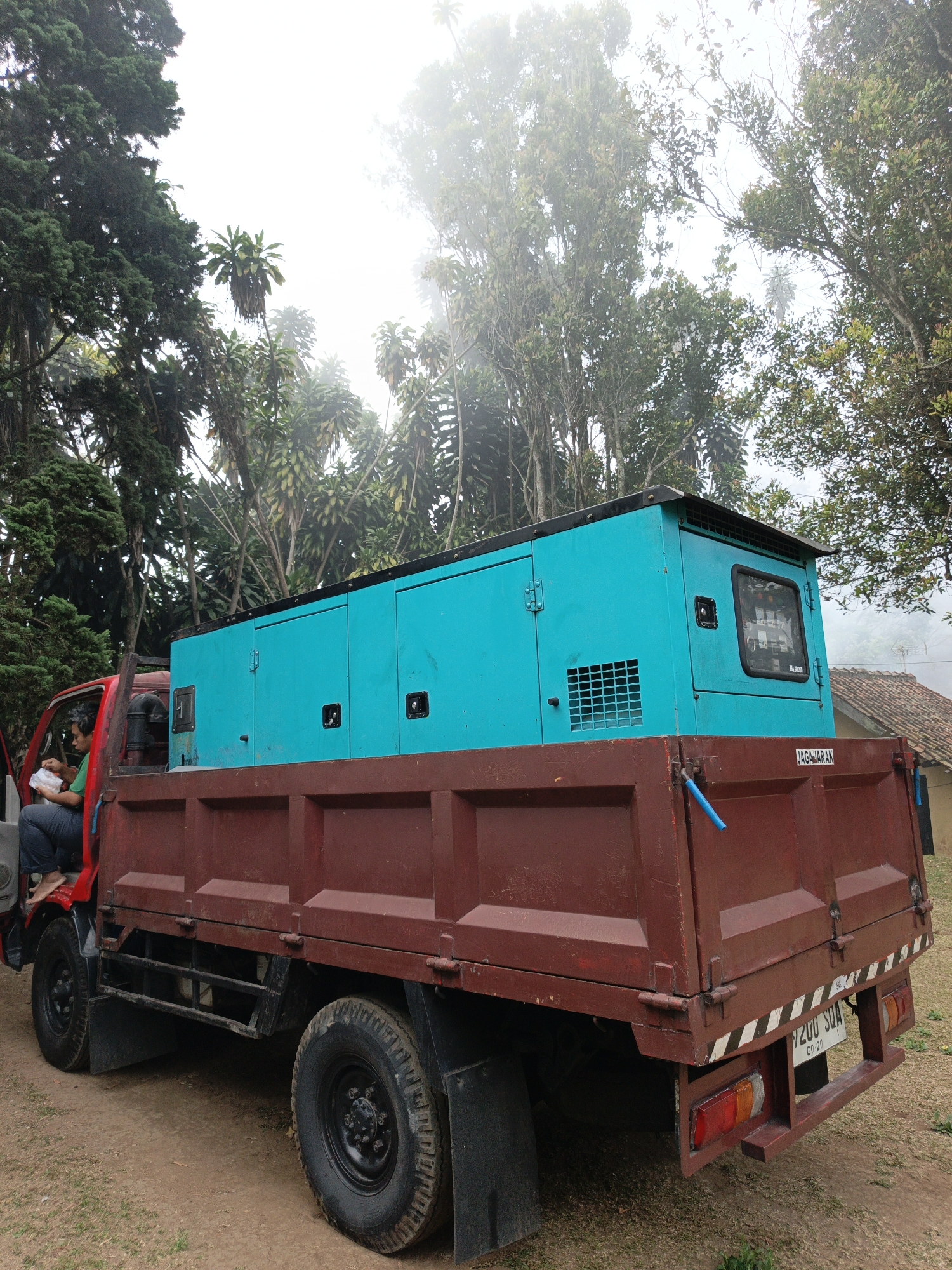 A side view of a red dump truck with a large, teal generator in its bed is parked on a dirt road. A man wearing a green shirt and dark pants sits in the driver's seat of the truck, holding a white plastic bag. The generator is a metal enclosure with ventilation grates and a small window. The truck has a brown bed and black tires. In the background, there are trees and a house with a tiled roof, suggesting a rural or semi-rural setting, possibly in Cibogo, Indonesia, given the license plate on the truck which reads "B 2100 SQA". The sky is overcast and hazy, with mist visible among the trees.