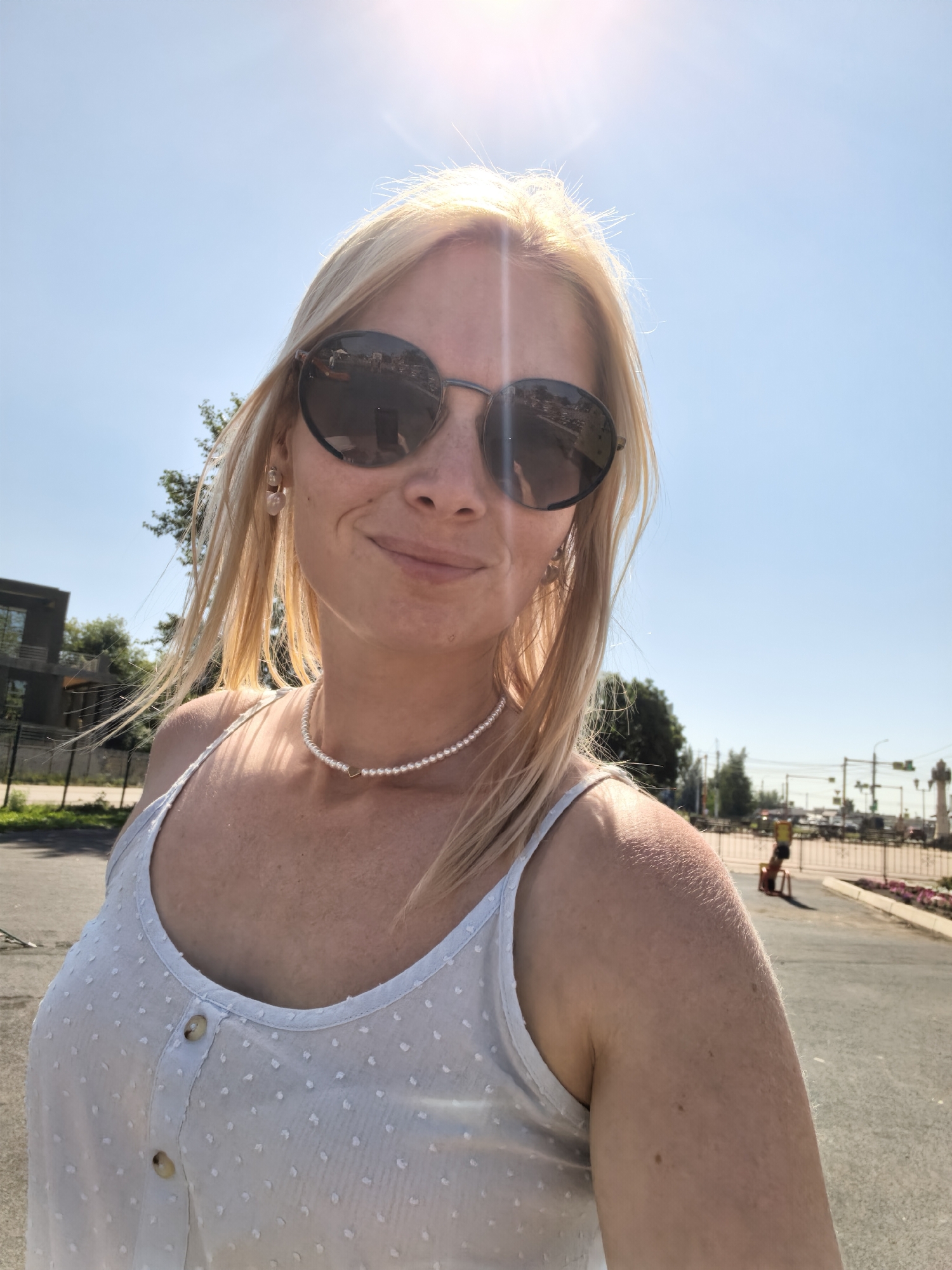 A blonde woman in her 20s wearing a light blue tank top, sunglasses, and a pearl necklace is taking a selfie outdoors on a sunny day. She is smiling and looking at the camera. Her hair is windblown. In the background, there is a clear blue sky, trees, buildings, and a street with traffic lights and cars. The image appears to have been taken on a street in the city of Sterlitamak, Russia.