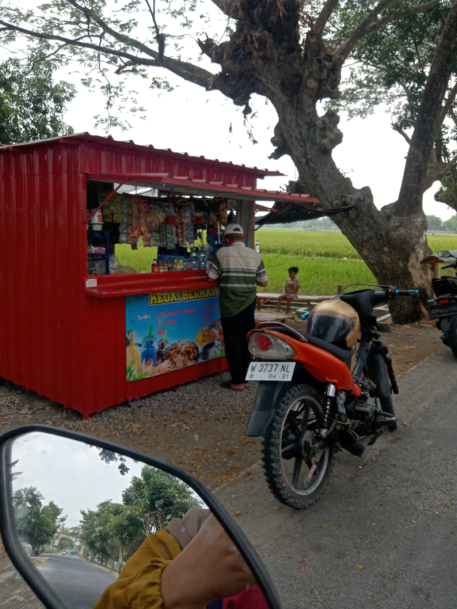The image depicts an outdoor scene in Mayangkawis, Indonesia, likely along a roadside or path. A bright red roadside kiosk, possibly a small shop or food stall, is prominently featured. Various packaged snacks and drinks are displayed on shelves within the open front of the kiosk. A sign on the kiosk reads "KEDAI BERKAH" and advertises "Sedia minuman sugar," "Es teh," and "Mie." A man in a green and white striped shirt and a white cap is standing by the kiosk, facing away from the camera. Further back, a young boy sits on a wooden bench in front of a lush green rice field, with a large, gnarled tree providing shade. In the foreground, an orange and black motorcycle is parked. A gold and black helmet rests on the motorcycle's seat. The license plate of the motorcycle reads "W 3737 NL 04-31." The reflection in the motorcycle's side mirror shows a road with trees and buildings. The ground around the kiosk is covered in gravel and scattered leaves.