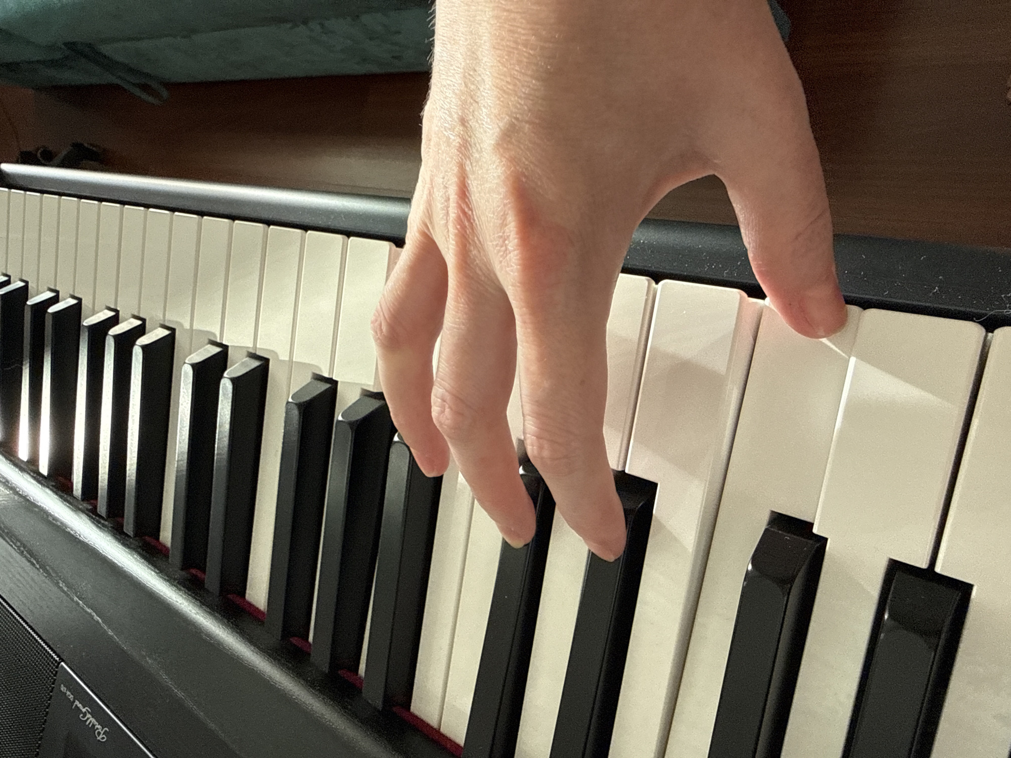 An overhead close-up shot captures a person's hand positioned over the black and white keys of a digital piano. The hand, with its fingers splayed slightly, is angled towards the right side of the frame. The piano keys are visible in a diagonal line, with black keys interspersed among the white keys. The background is softly blurred, suggesting an indoor setting, possibly a room in a home, with a hint of a dark wooden floor and a teal-colored object, perhaps furniture, on the right. The lighting in the image appears to be natural, casting subtle shadows on the piano keys and the hand. The piano itself is black, with some branding or text visible on the upper left corner of the piano's surface, which appears to read "Roland."