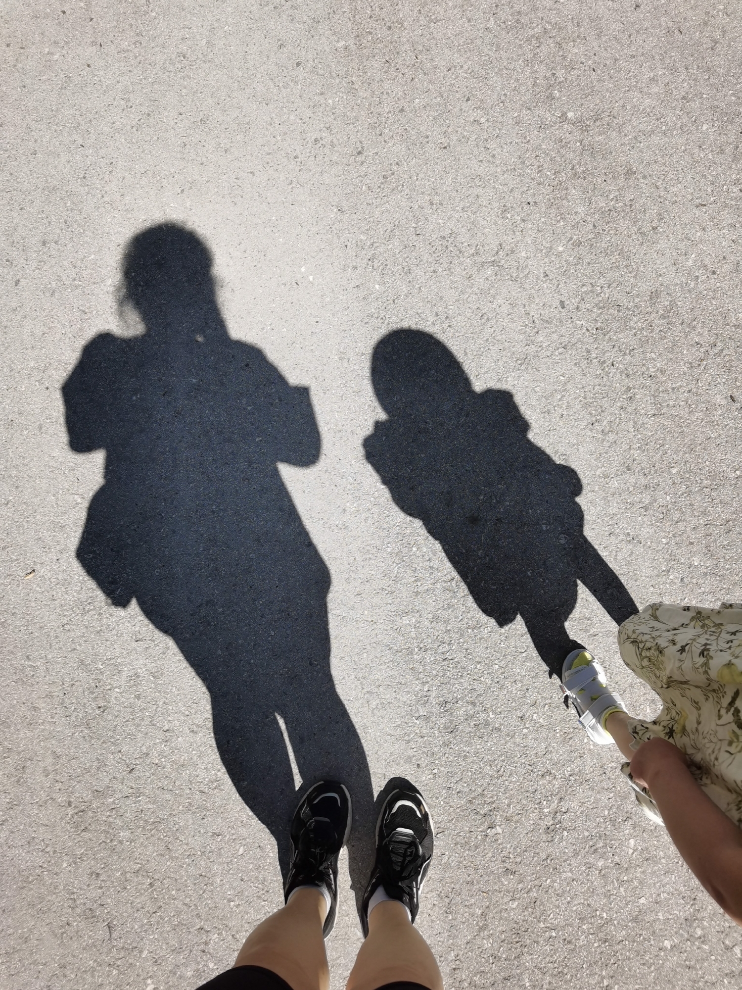 The image shows a top-down view of a light-colored asphalt surface with shadows of two people. One shadow appears to be of an adult wearing a jacket, while the other is of a child holding the adult's hand. The lower part of the image shows the legs and feet of the person taking the picture, wearing black running shoes and shorts. To the right, the feet of the child are visible, wearing a white and yellow orthopedic shoe and a dress. The location and details of the city are not present in the image.