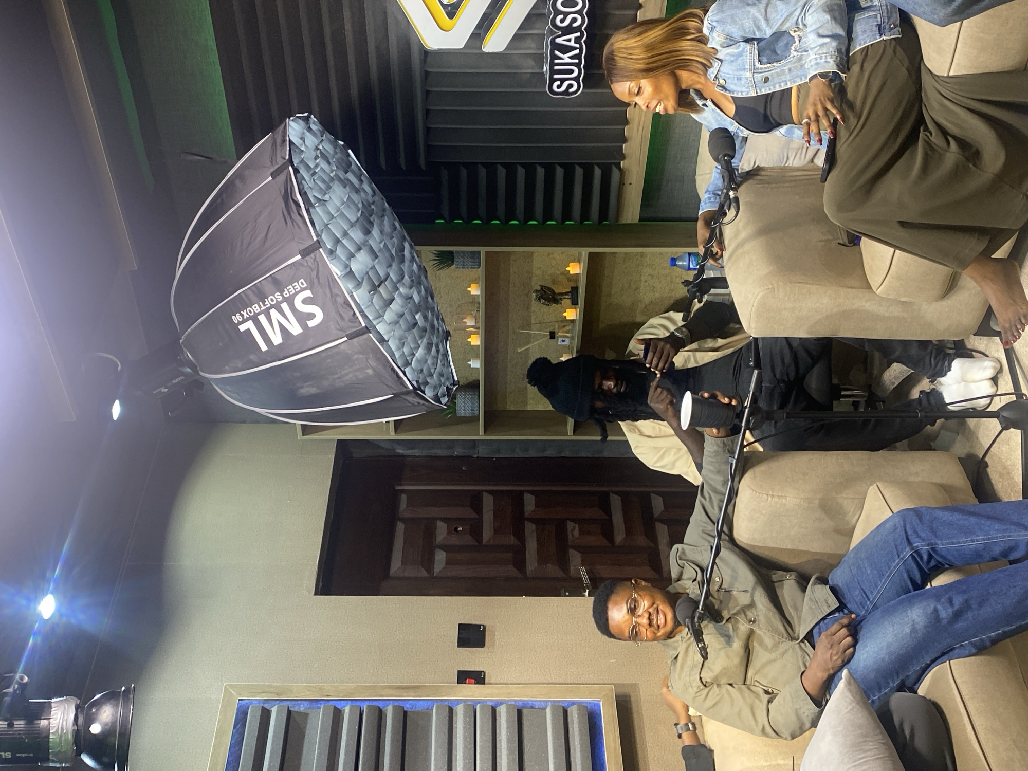 The image depicts a podcast recording session in a studio in Gura Top, Nigeria. Three individuals are seated on a beige sectional couch. On the left, a man wearing glasses, a green jacket, and blue jeans smiles and looks towards the camera. In the center, a man with dreadlocks, a black beanie, and a tan shirt is holding a phone and a white cup. On the right, a woman with shoulder-length brown hair, a black crop top, and a denim jacket is speaking and looking to her left. She is wearing a long, olive-green skirt. The studio environment features soundproofing panels on the walls, shelves with decorative items and candles, a large softbox light suspended from the ceiling, microphones, and studio equipment. A prominent "SUKA SO" logo is visible on a wall in the background.