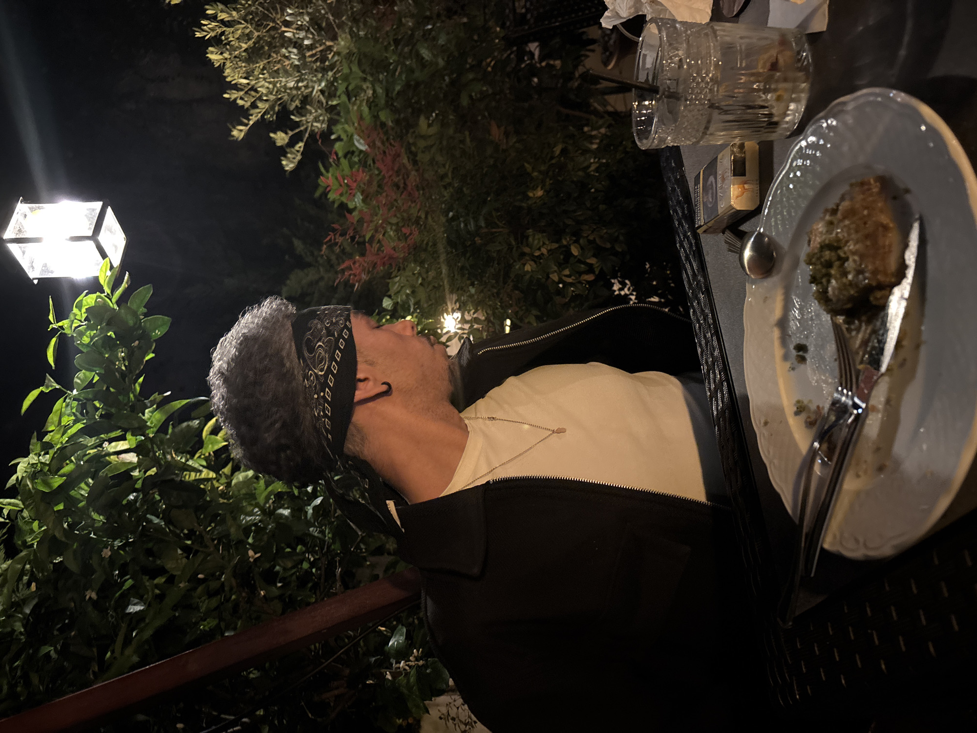 A person with gray hair and a paisley bandana is seated at a table at an outdoor restaurant. The person is wearing glasses, a white t-shirt, and a black jacket. A plate of food, a glass of water, a spoon, a fork, and a pack of cigarettes are on the table. The restaurant appears to be in Lisbon, Portugal, as indicated by the type of plants and the architecture. A light fixture is visible in the background. The scene is at night.