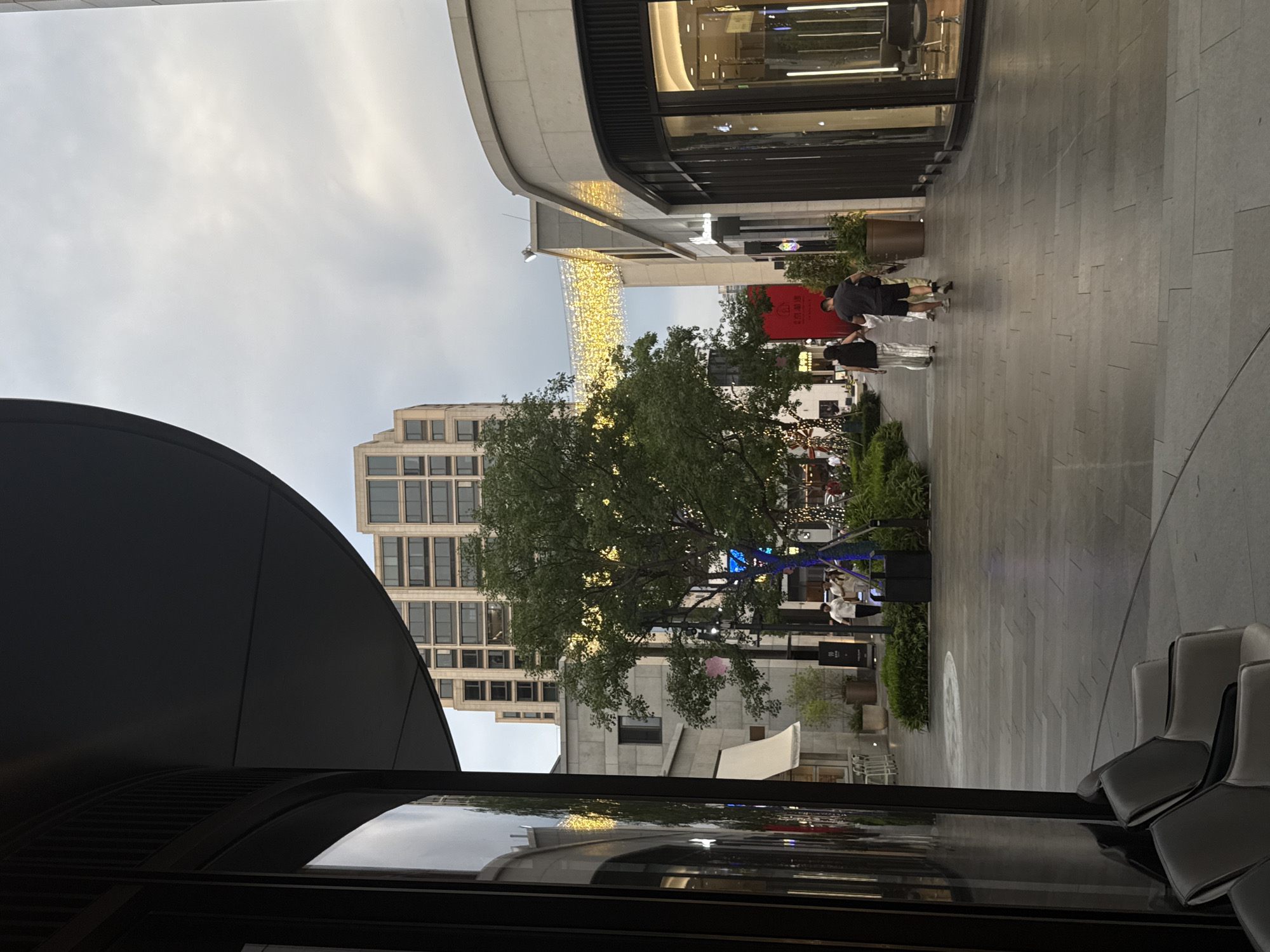 The image depicts a pedestrian walkway in Yiwu, China, likely a commercial district, with modern architecture and outdoor seating. In the foreground, the curved facade of a building with large windows and dark trim is visible on the left. To the right of this, the walkway is paved with large, rectangular gray tiles. A row of modern, minimalist chairs with light-colored upholstery and dark frames is arranged along the left edge of the walkway.
Further down the walkway, a large, mature tree with lush green foliage is illuminated by numerous small, warm-toned lights, and a string of bright yellow decorative lights spans across the walkway overhead. Several people are walking away from the viewer: a couple in white tops and striped pants, followed by a man in a dark t-shirt and shorts. They are walking on the right side of the walkway. In the background, a tall, multi-story building with a grid-like facade of windows stands prominently. Other buildings with stone exteriors and shops with large glass storefronts line the walkway. The sky is overcast with a diffused light, suggesting it is late afternoon or early evening. Planters with greenery are placed along the sides of the walkway. The overall impression is of a well-maintained, contemporary urban public space.