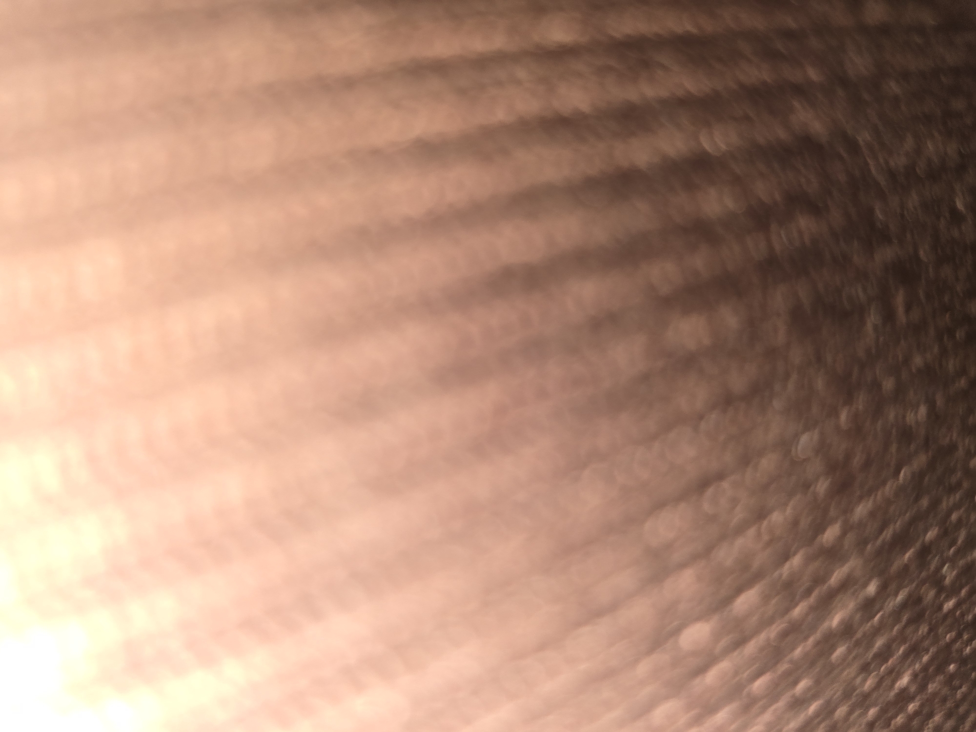 This is a close-up, abstract photograph of a textured surface, possibly fabric or a metallic material, with a warm, diffused light source from the upper left. The texture appears to be a series of diagonal ridges or lines, creating a sense of depth and movement. The color palette is dominated by shades of peach, tan, and brown, with lighter highlights where the light hits most intensely. There are no discernible people, animals, or specific objects in the image, nor is there any indication of a specific location within the city of Ulan-Ude, Russia. The image is abstract and focuses on light, texture, and color.