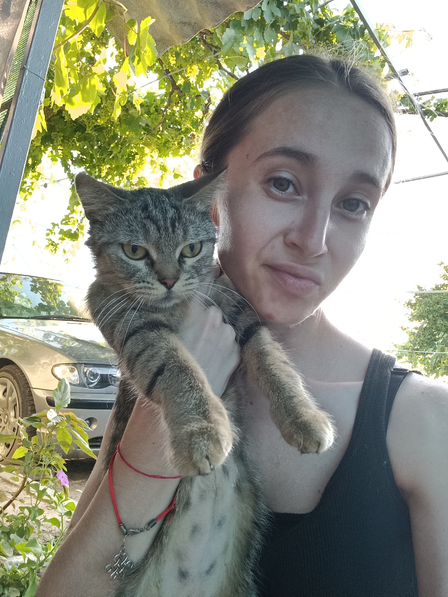 In Vypasne, Ukraine, a young woman with light brown hair, pulled back into a bun, is pictured outdoors, holding a tabby cat. The woman is wearing a black tank top and a red string bracelet with a silver charm. The cat, held horizontally across her chest, has grey and black stripes and green eyes. In the background, a silver car is parked, and green foliage and grapevines are visible, suggesting a rural or semi-rural setting. The lighting indicates it is daytime, with the sun casting bright light through the leaves.